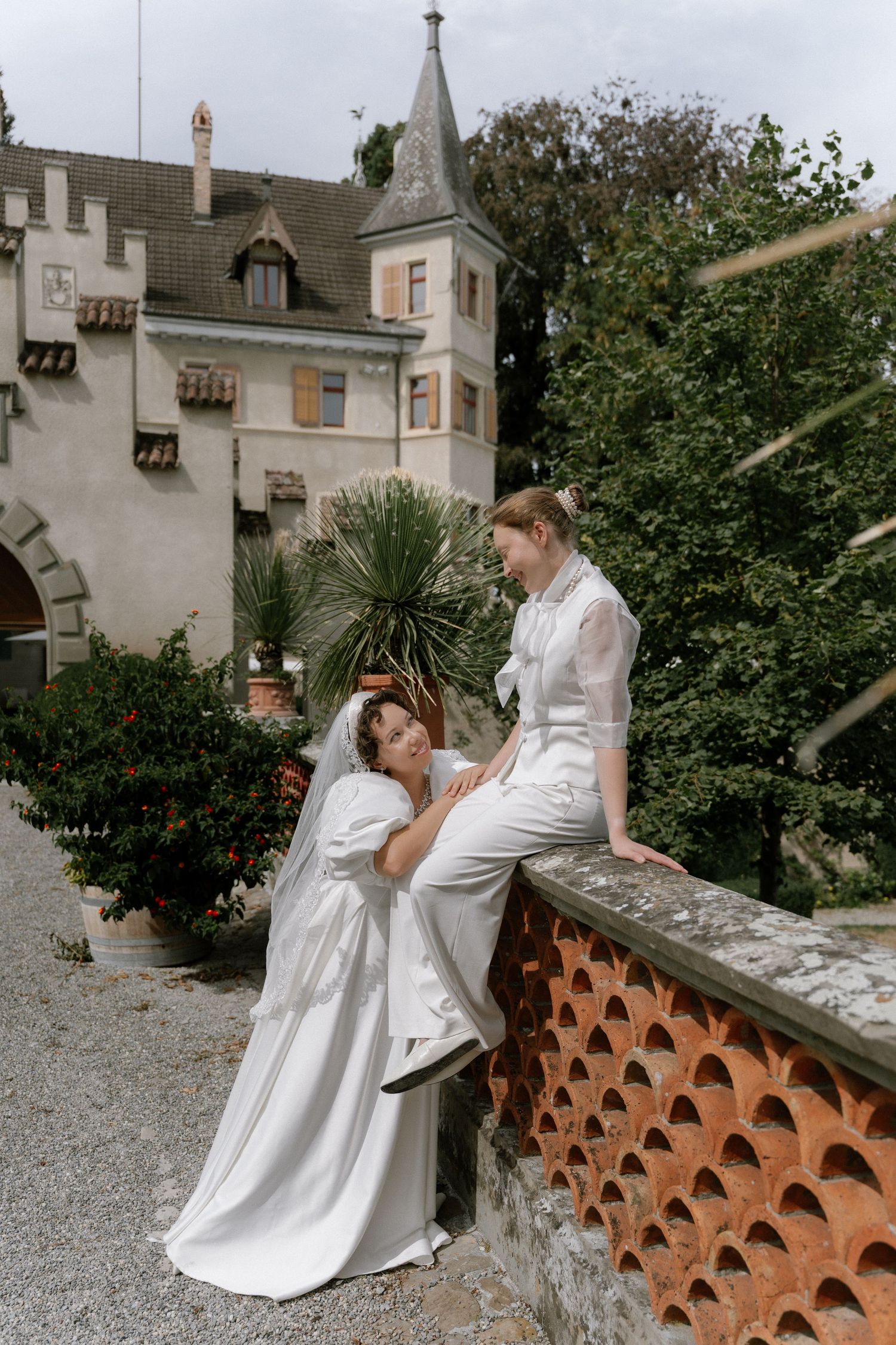 Two brides in white wedding attire pose together outside a European castle venue surrounded by lush greenery.