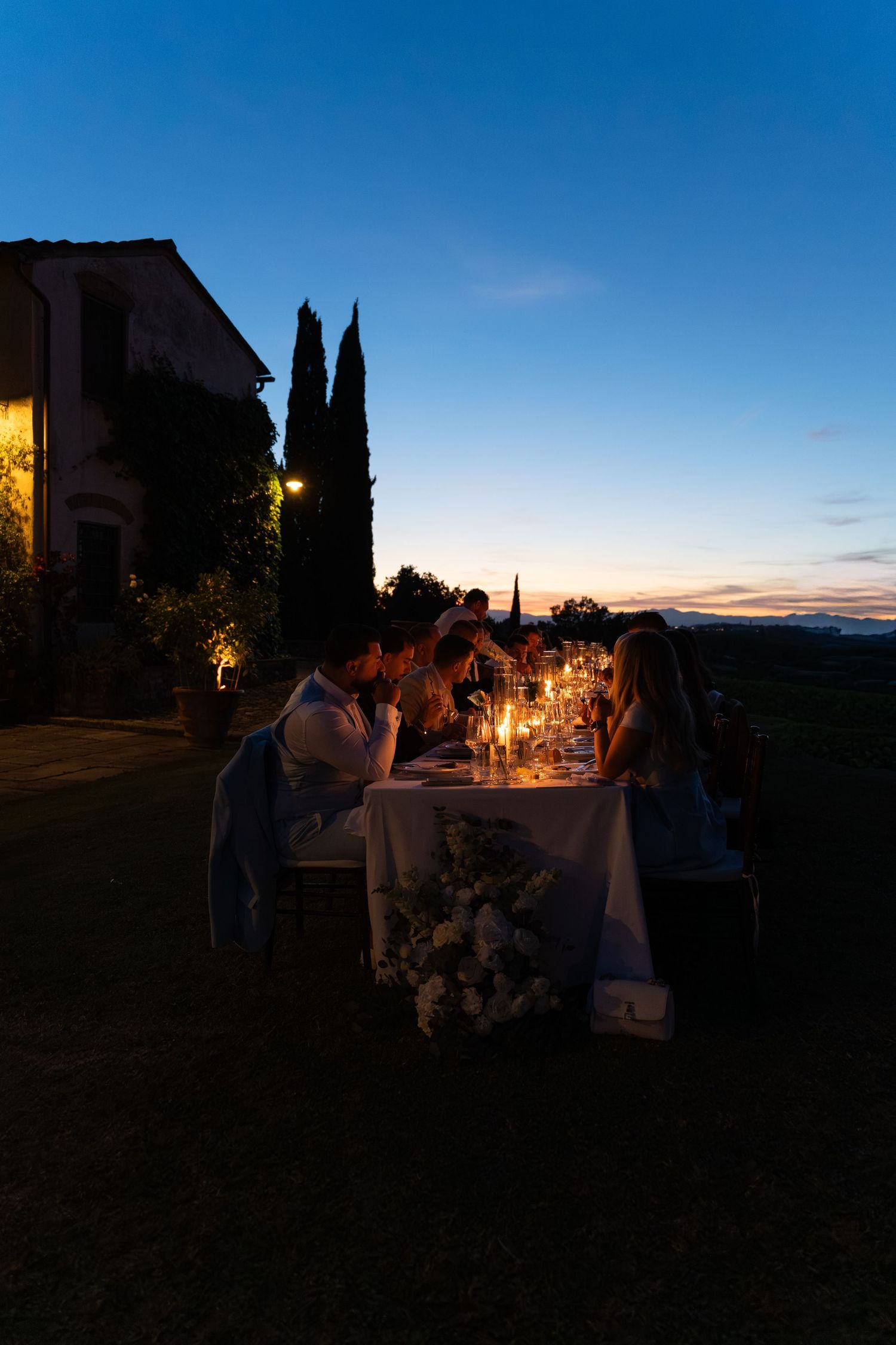 Guests dine by candlelight at an outdoor table during a stunning Tuscan sunset, surrounded by cypress trees and rolling countryside.