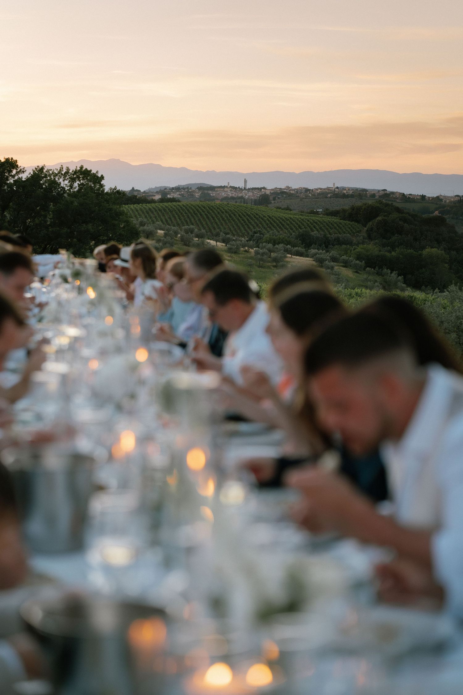 Guests dining at a long outdoor table during sunset with vineyard views and candlelight in a scenic countryside setting.