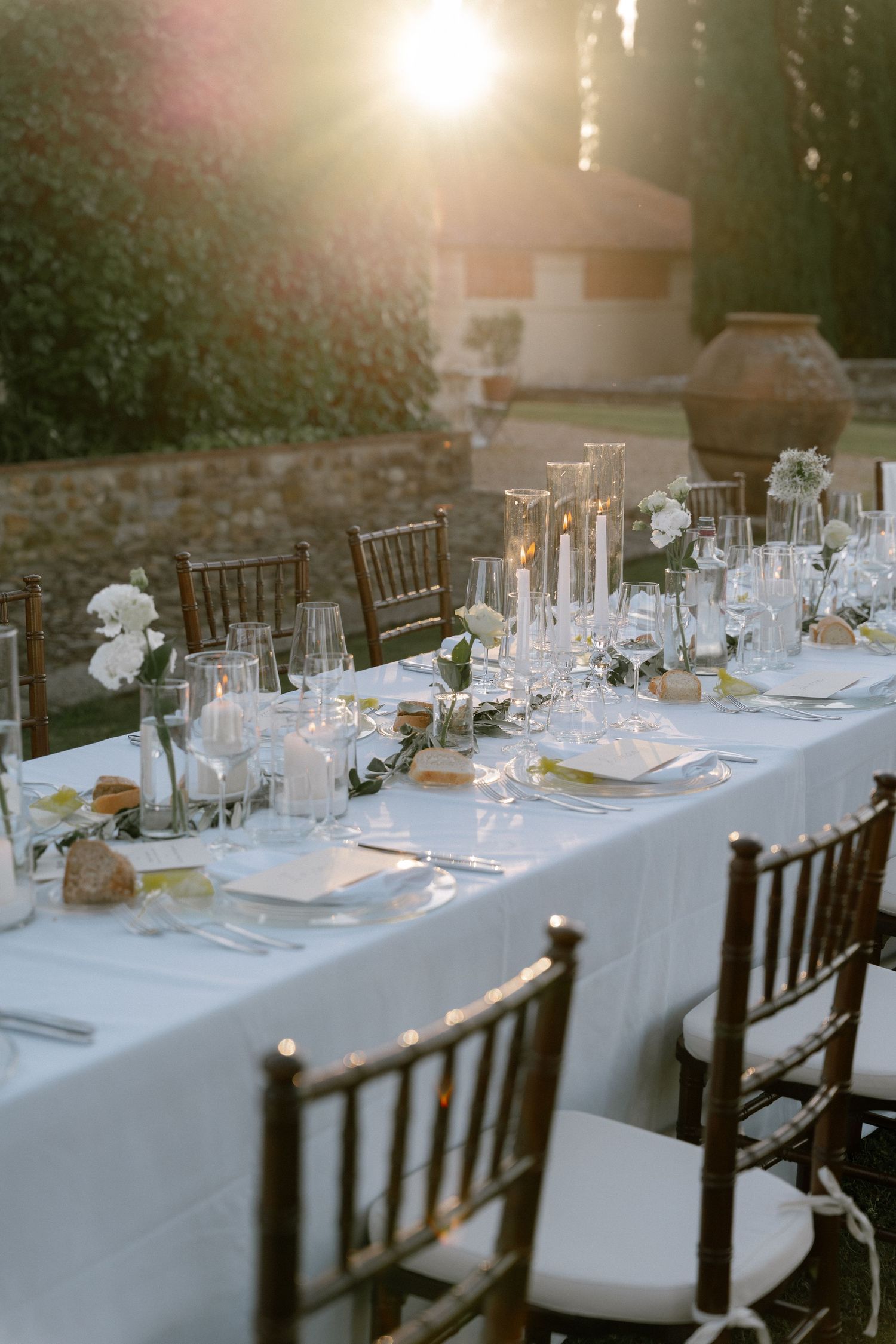 Elegant outdoor wedding reception table set with white linens, candles, and floral arrangements at golden hour.