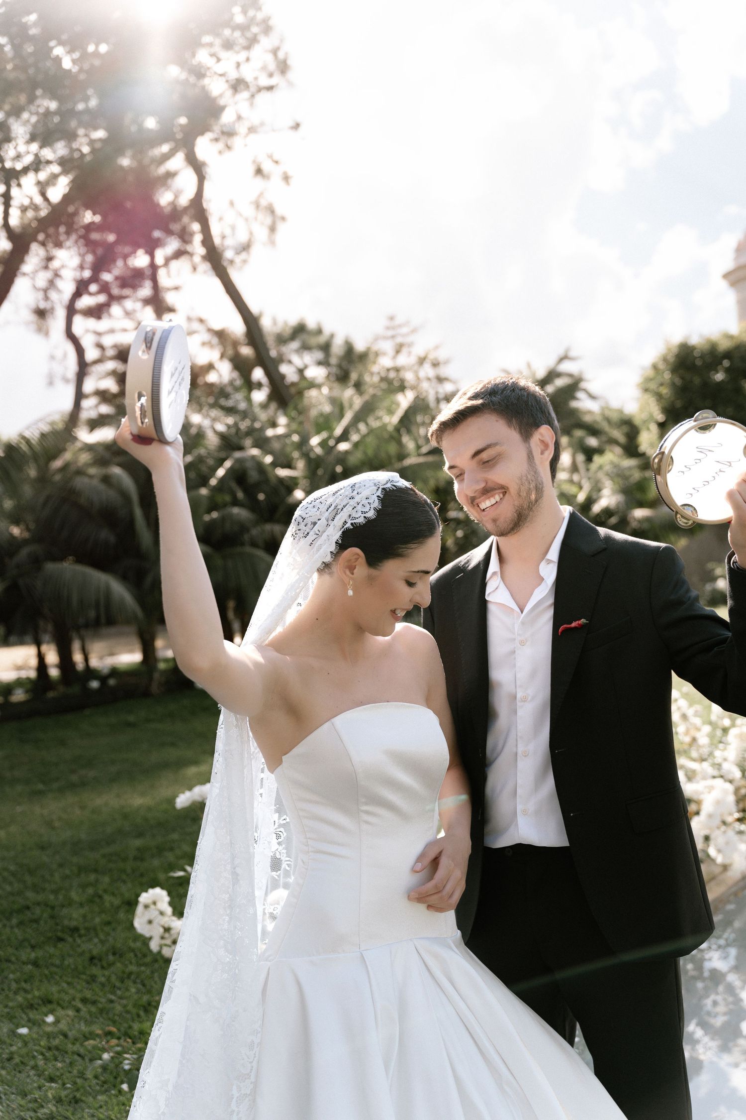 A joyful bride in a white strapless gown and veil dances with a smiling groom in a black suit outdoors.