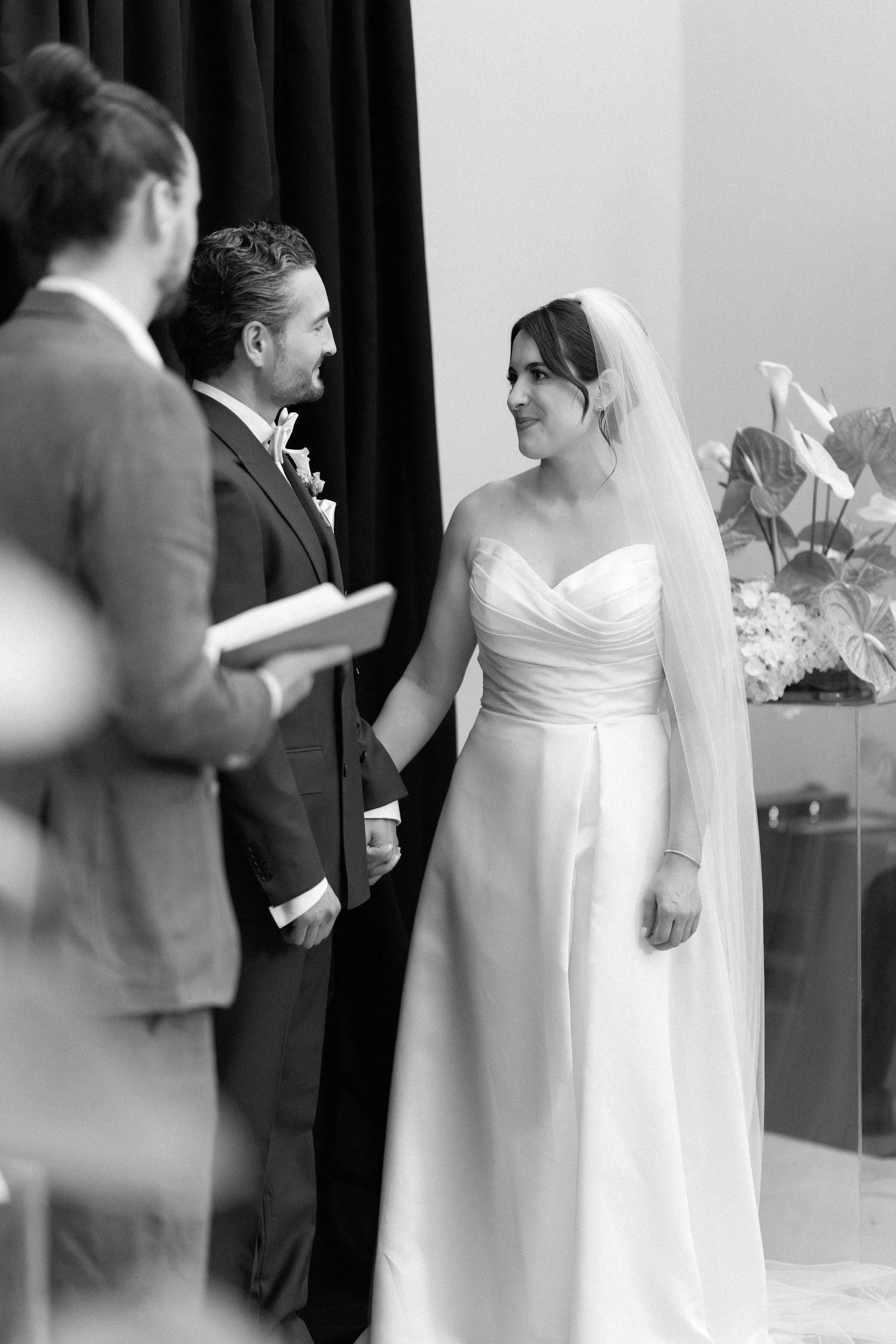 Black and white photo of a bride and groom holding hands during their wedding ceremony with an officiant.