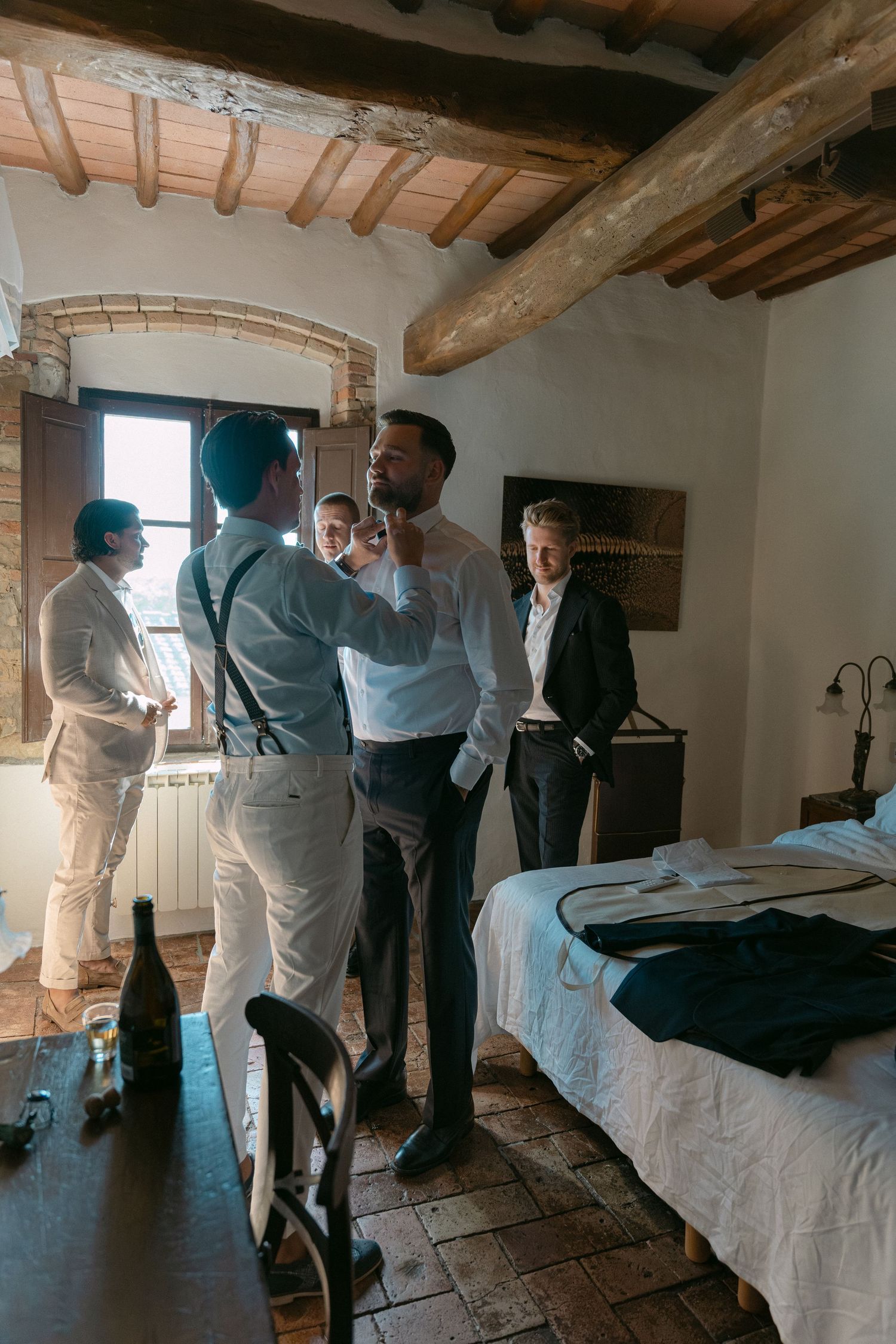 Groomsmen helping each other get dressed in a rustic Tuscan villa room before a wedding ceremony.