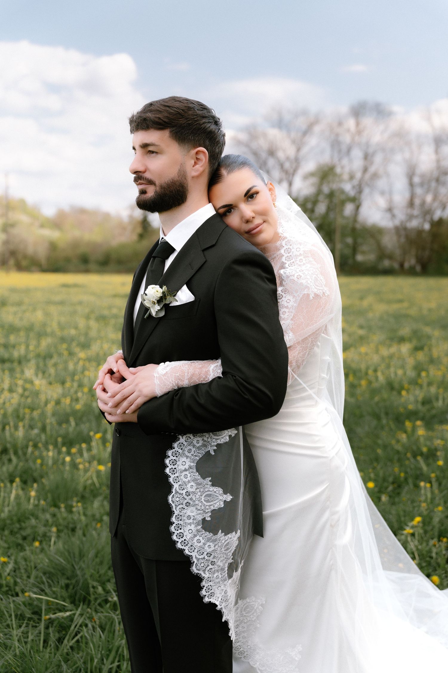 Bride in white lace veil embraces groom in black tuxedo outdoors in a green field with yellow flowers.