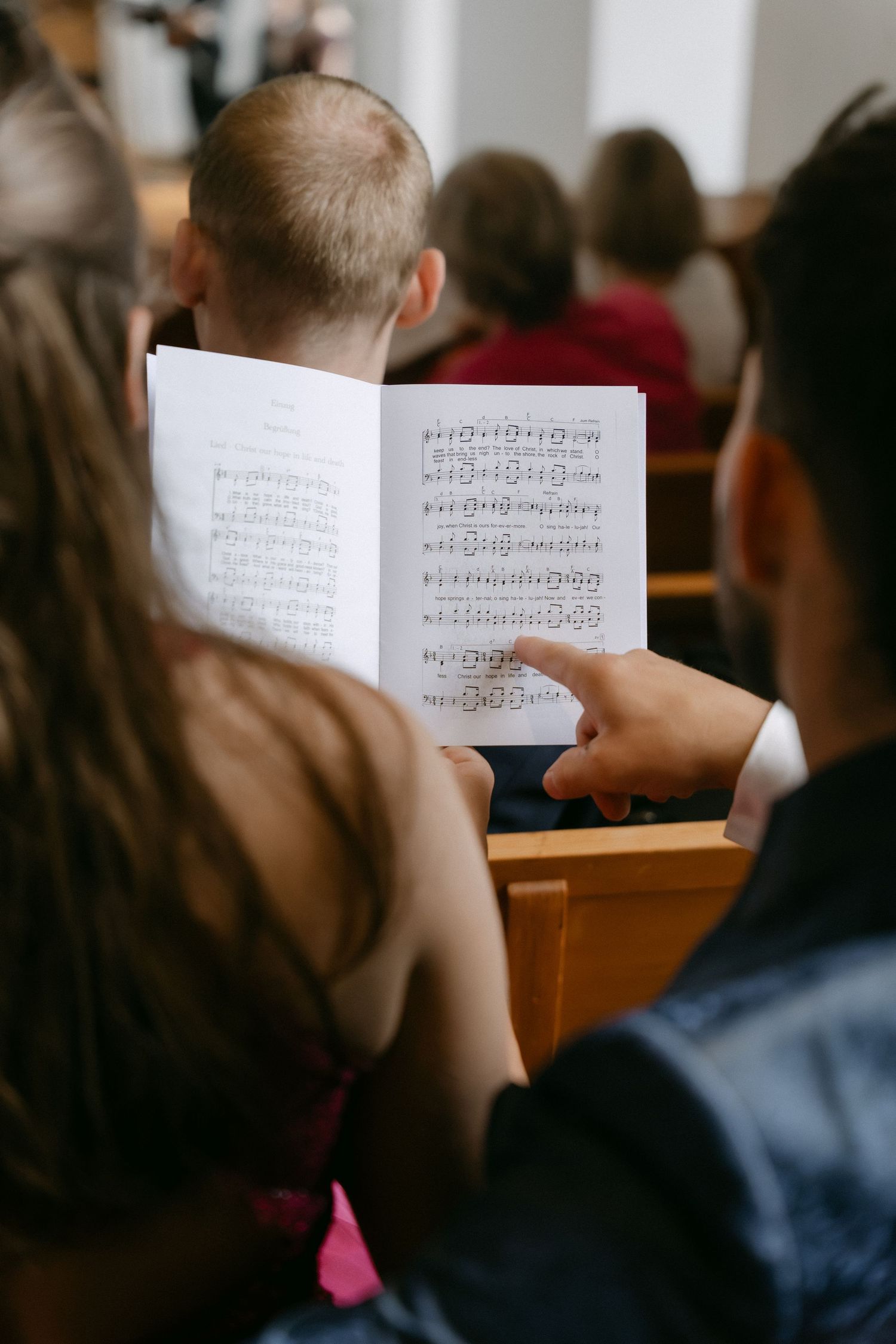 Congregation members seated in church pews share an open hymnal with printed sheet music while a man points to the notes.