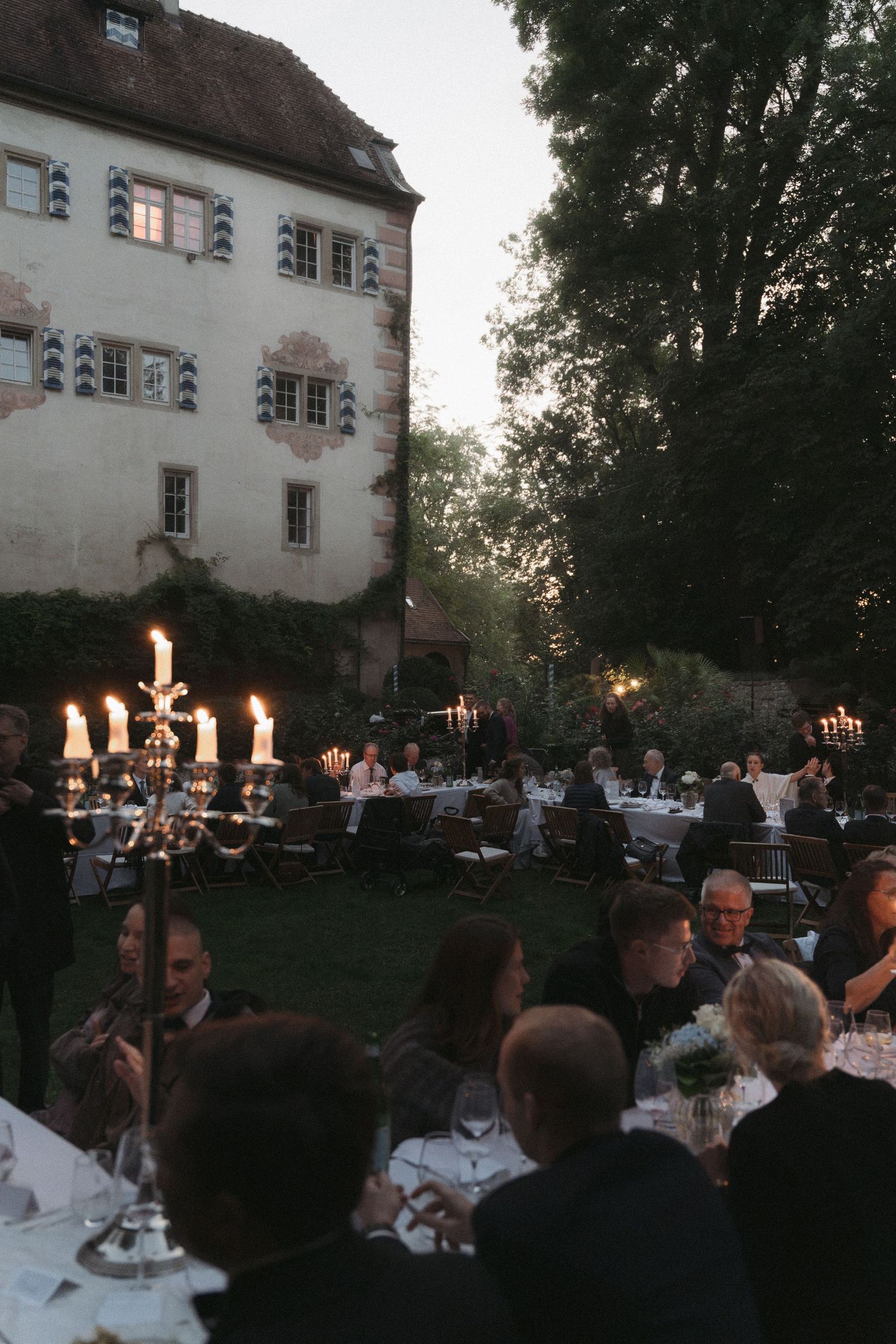 Guests dine at long tables on a lawn at dusk beside a historic building, lit by a candelabra in the foreground.