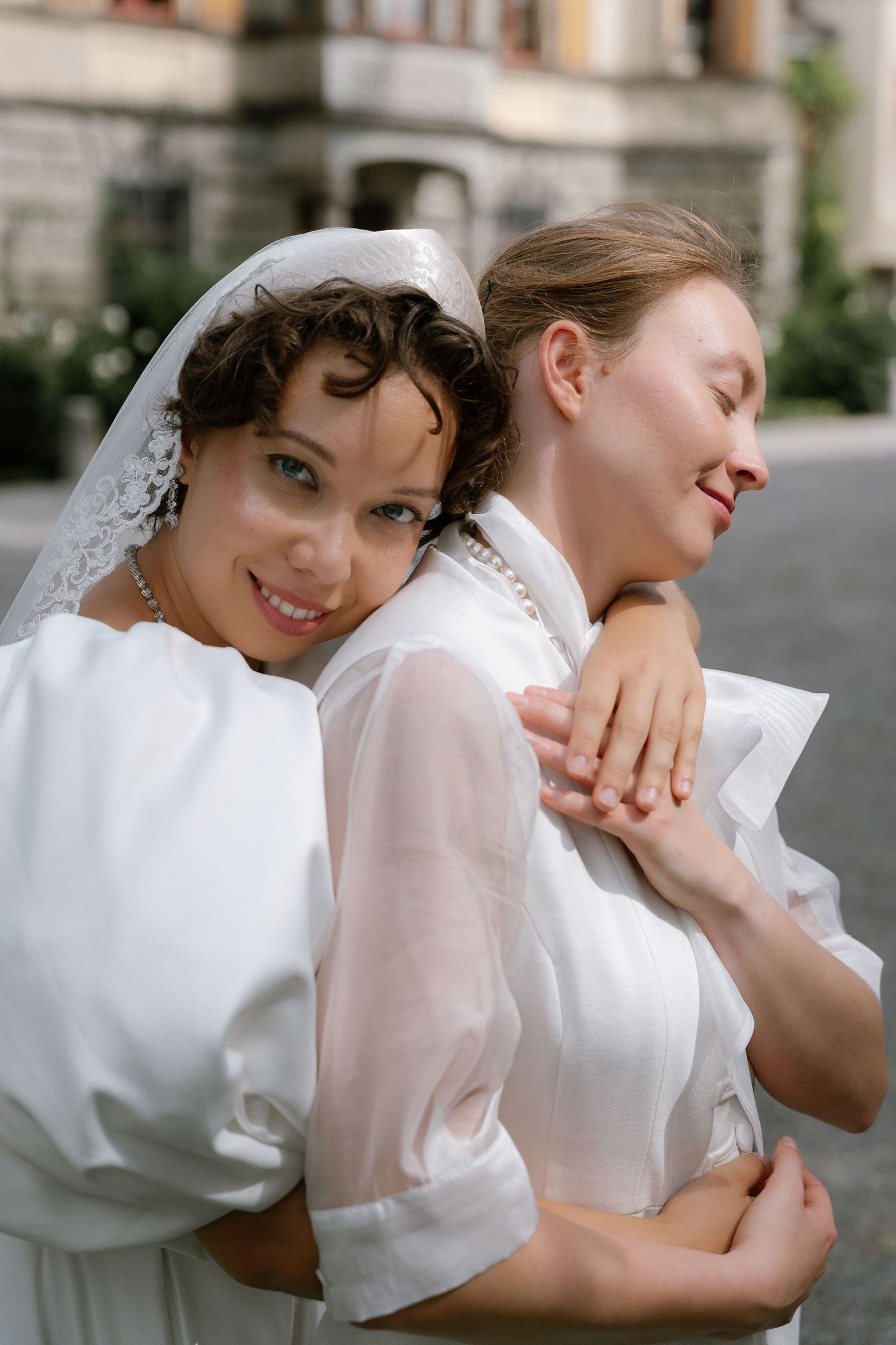 Two brides in white dresses embrace outdoors, smiling joyfully on their wedding day.