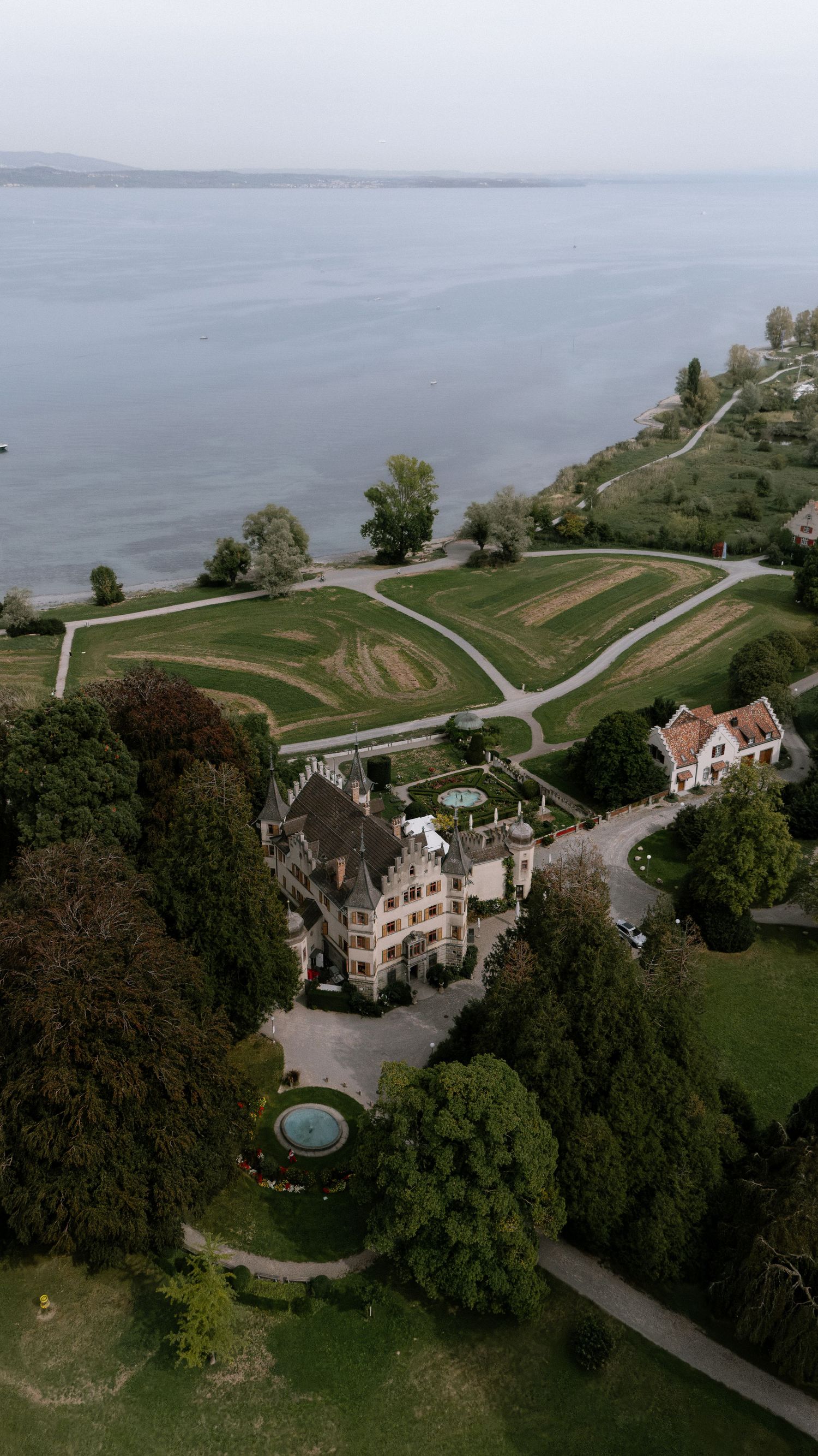 Aerial view of a grand estate with manicured gardens and fountain, surrounded by trees beside a serene lake.