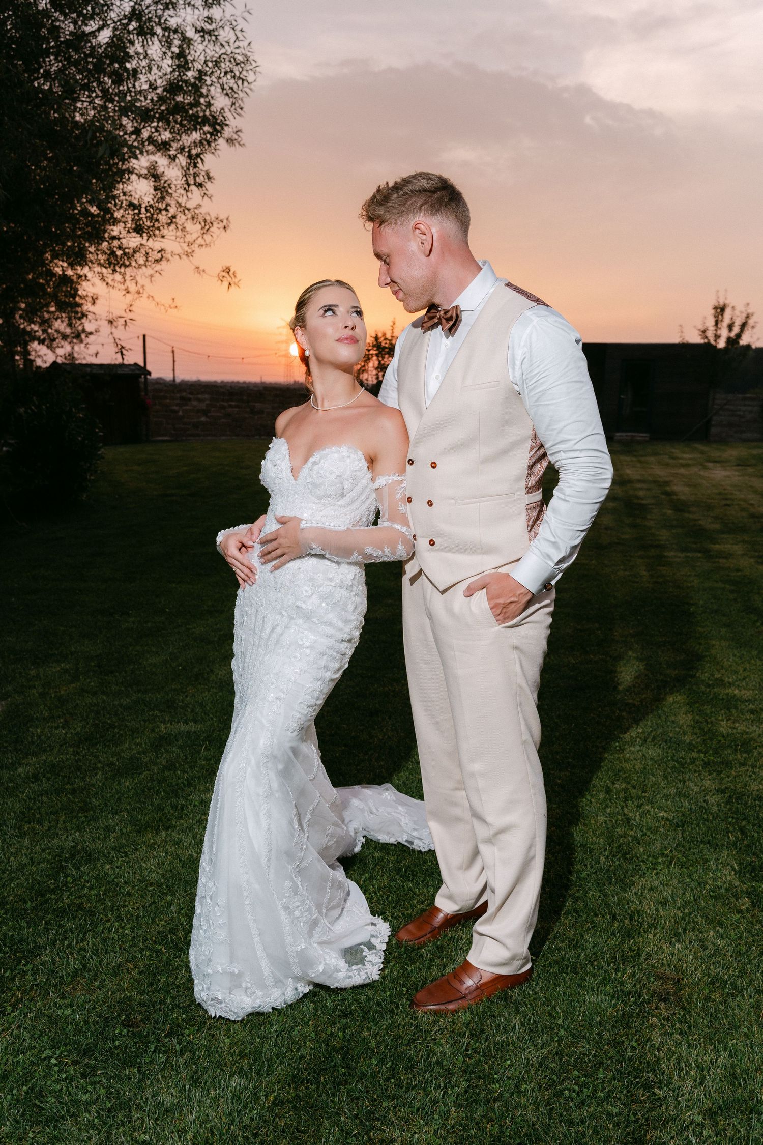 A couple shares a romantic moment at sunset on their wedding day, wearing elegant white and cream formal attire.