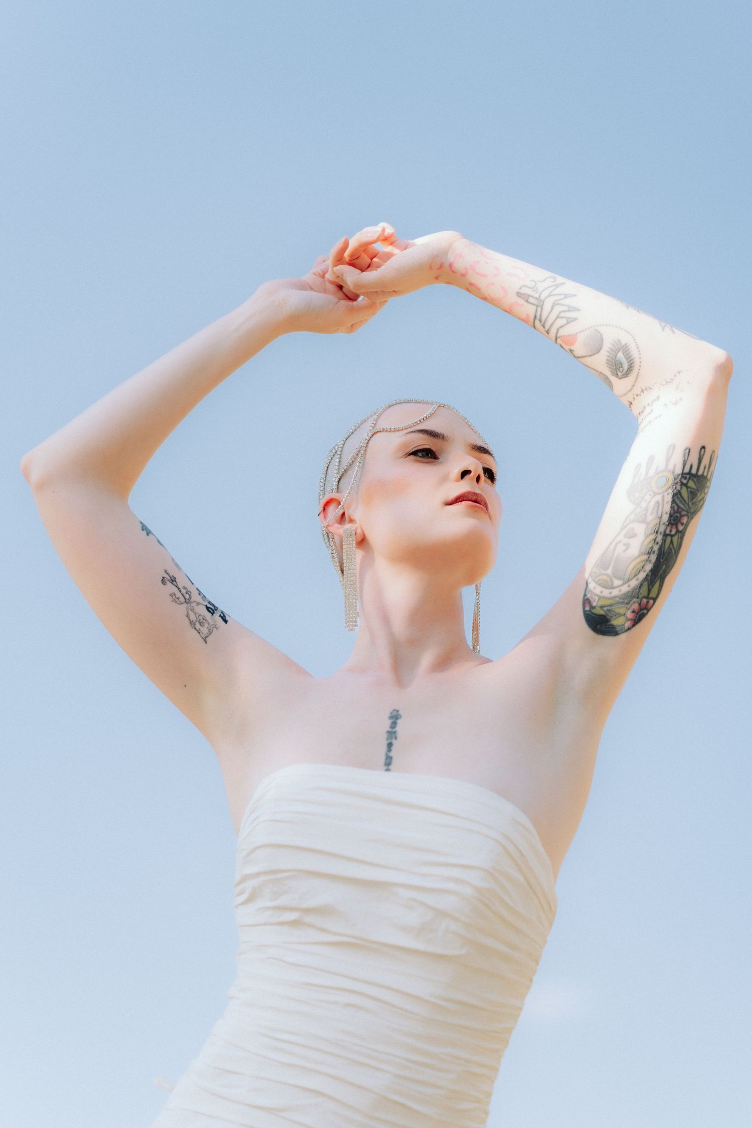 Person in white strapless dress poses gracefully against blue sky background, showcasing delicate arm movements.