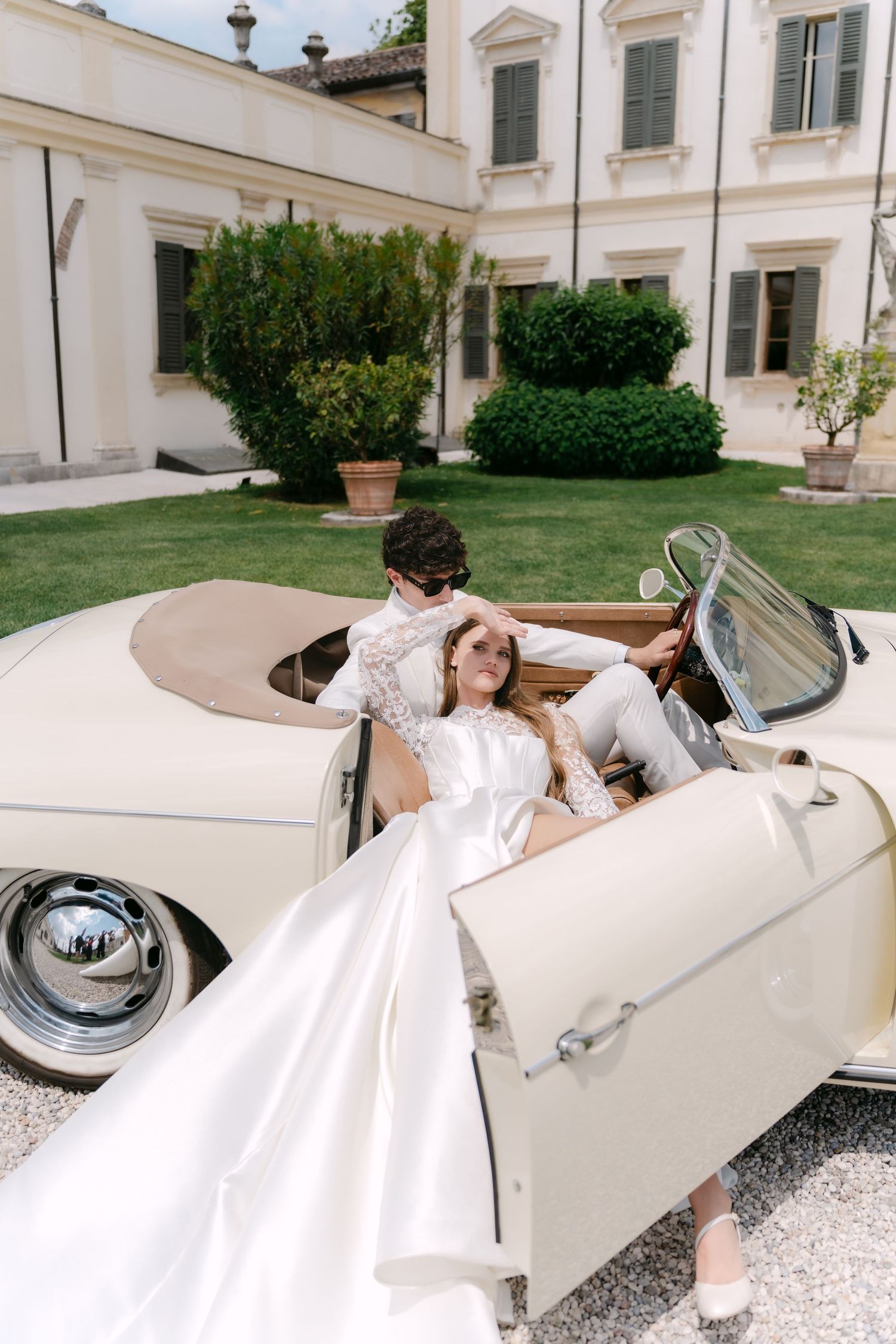 A bride in a flowing white dress poses playfully on a vintage cream-colored car outside an elegant white building. Villa Mosconi Bertani
