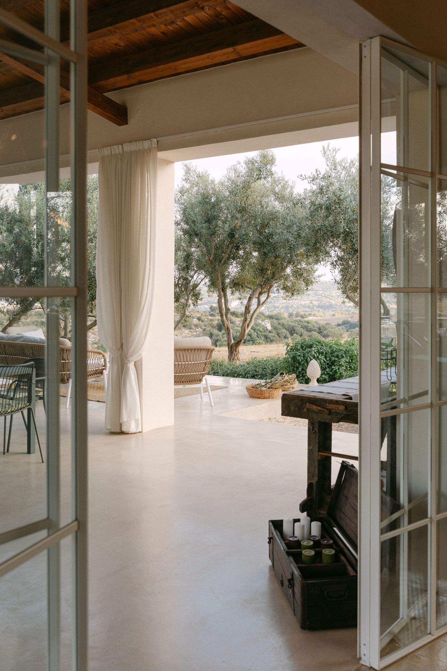 Bright Mediterranean patio with white curtains, exposed wooden beams, and olive trees visible through French doors.