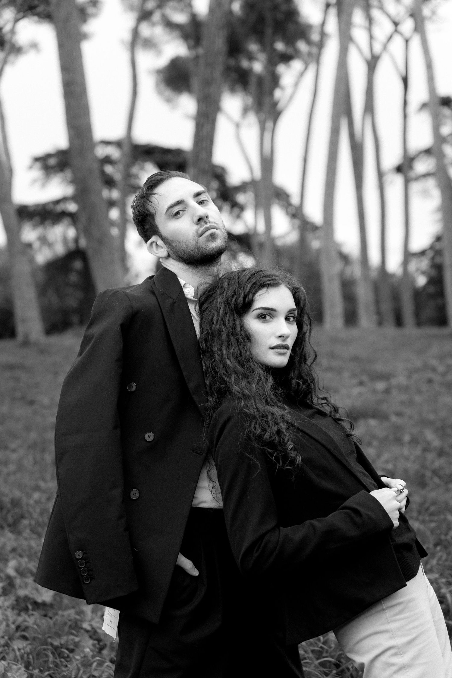 Moody black and white fashion photograph of a couple in dark coats posed against a forest backdrop.