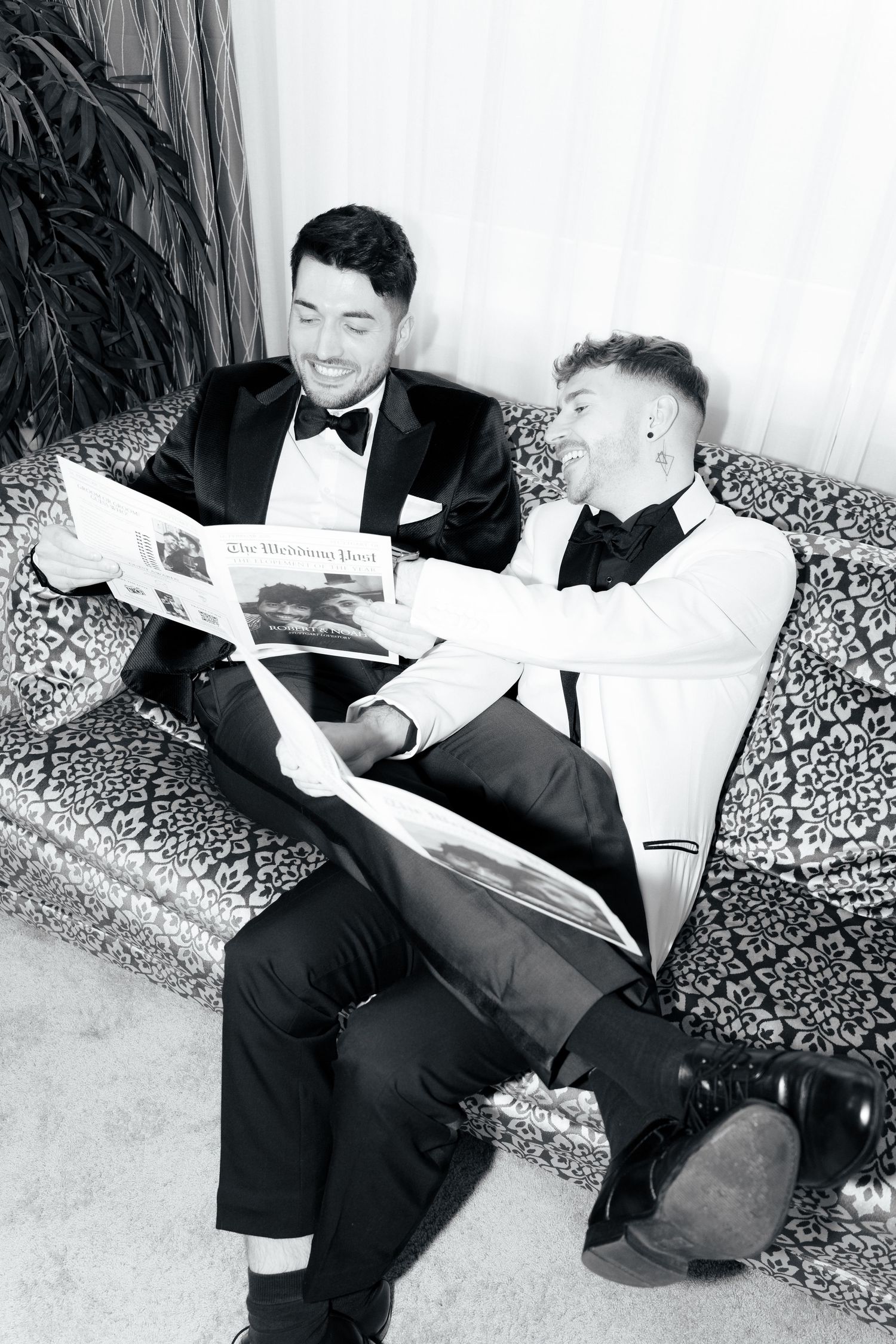 Two men in formal attire sit on a patterned couch reading documents together in black and white photograph.