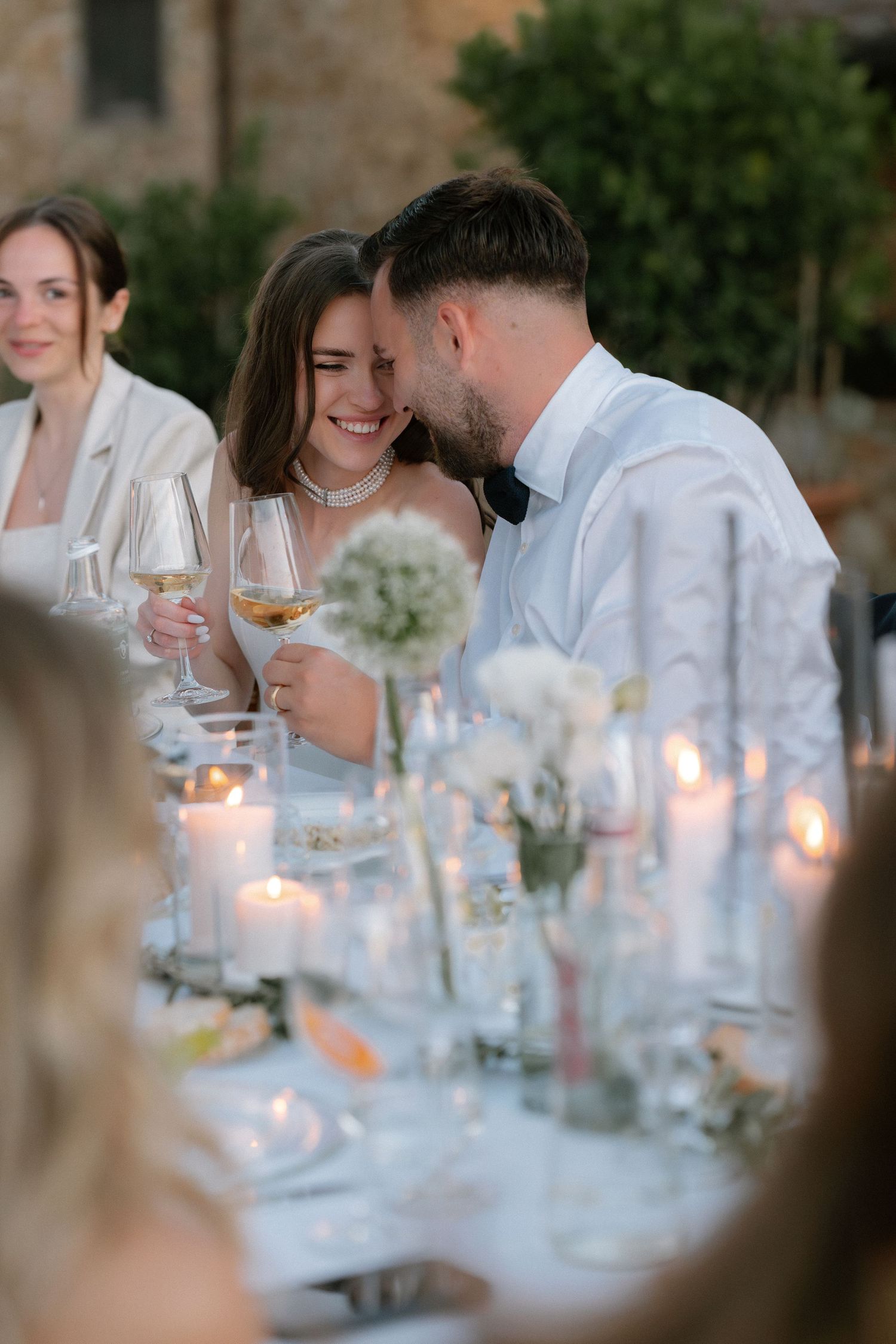 Bride and groom share an intimate moment at candlelit outdoor wedding reception table, holding white wine glasses.
