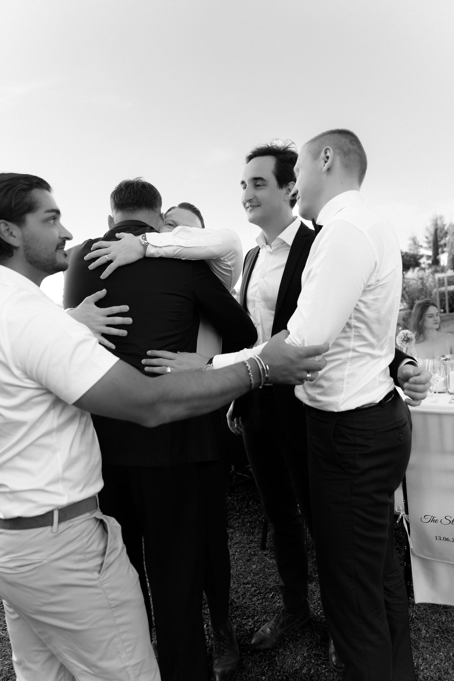 Black and white photo of groomsmen sharing a group hug at an outdoor wedding reception.