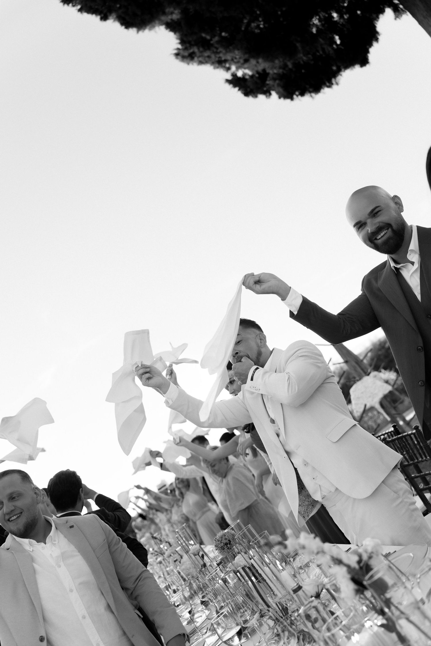 Black and white photo of smiling men in suits waving white napkins at an outdoor celebration event.