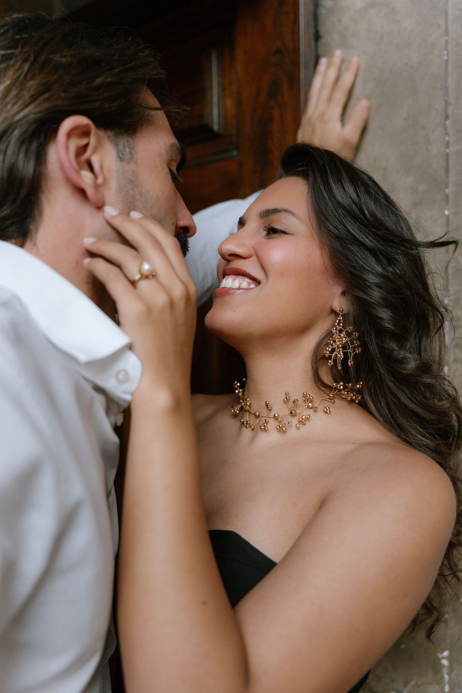 A smiling woman in gold jewelry and black dress gazes at a man in white shirt near a wooden door.