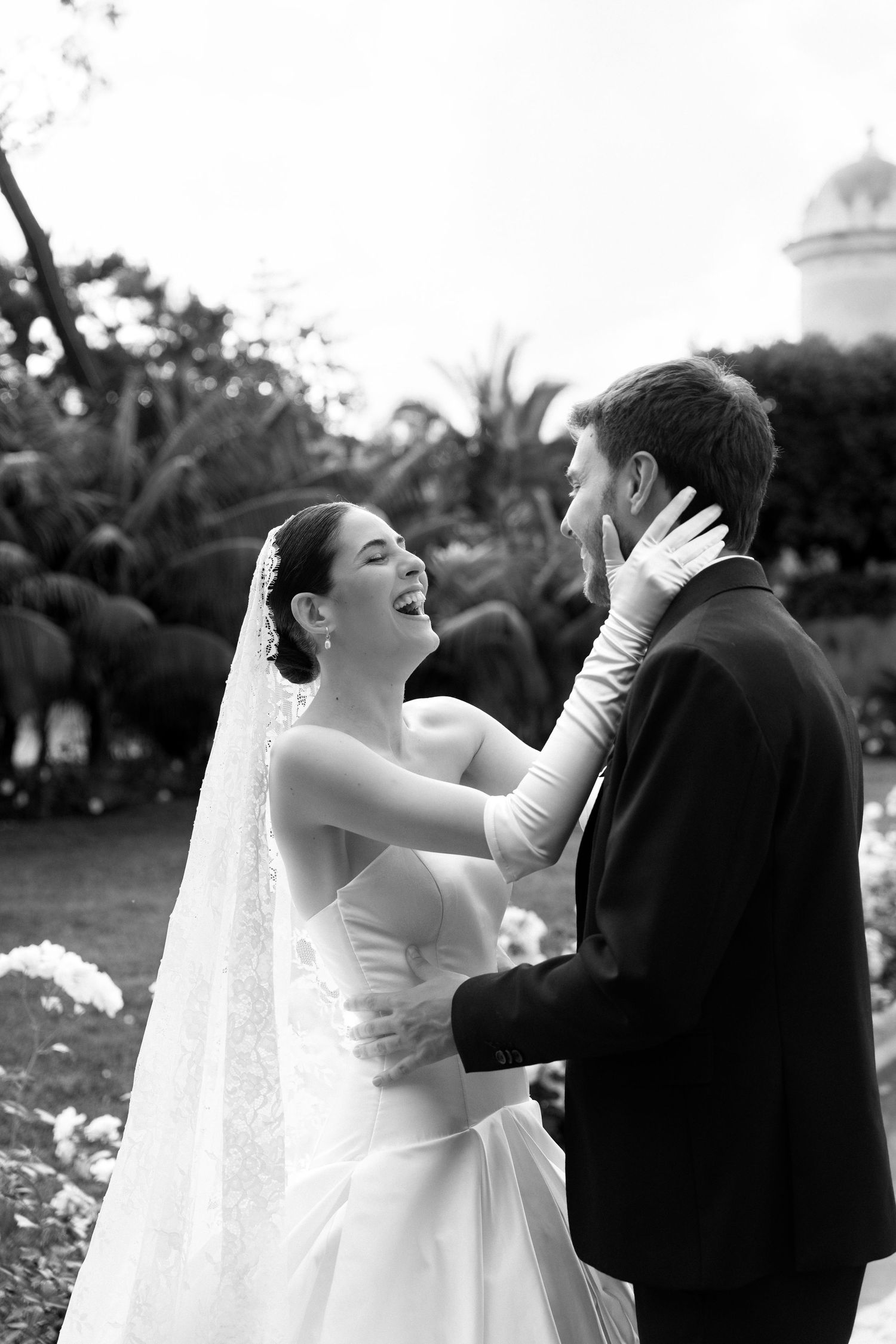 Black and white photo of a laughing bride in veil and gloves tenderly touching grooms face in a garden setting.