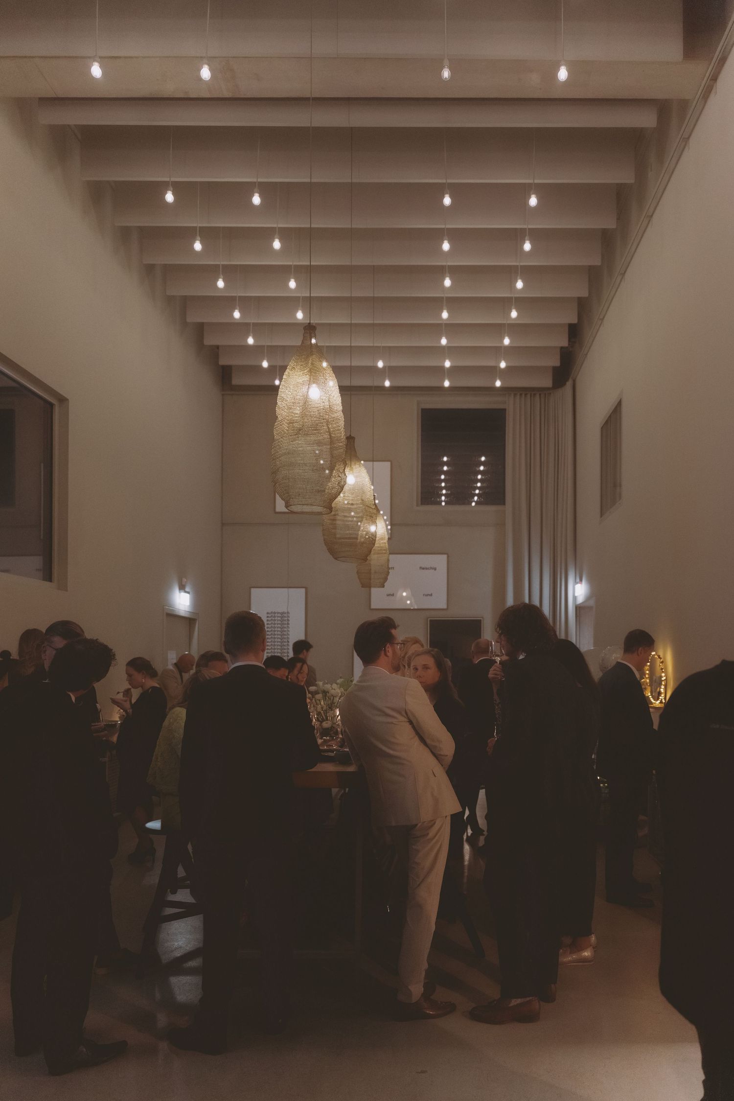 Elegant indoor gathering with guests mingling under decorative pendant lights and twinkling ceiling bulbs in a warmly lit venue.