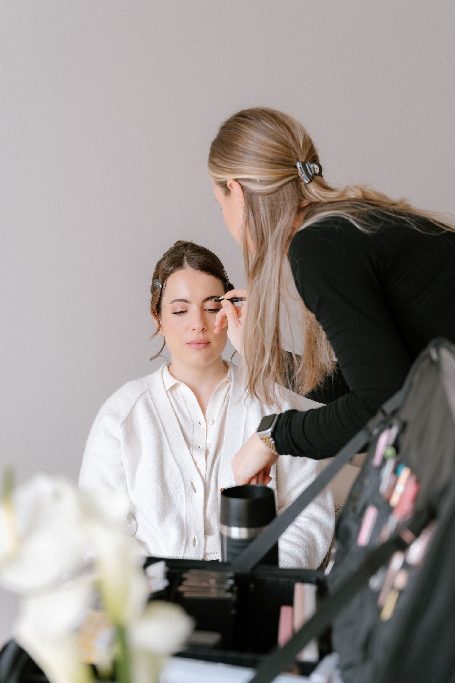 Professional makeup artist applying eye makeup to a young brunette woman in a bright studio setting.