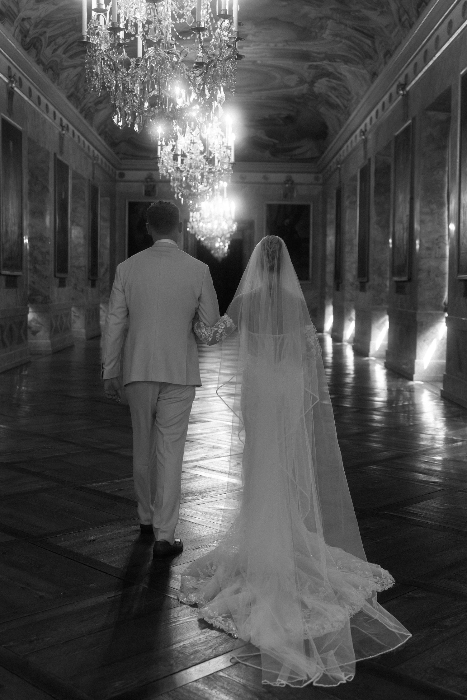 A couple walks through an ornate palace ballroom with crystal chandeliers and polished floors in black and white. Schloss Ludwigsburg