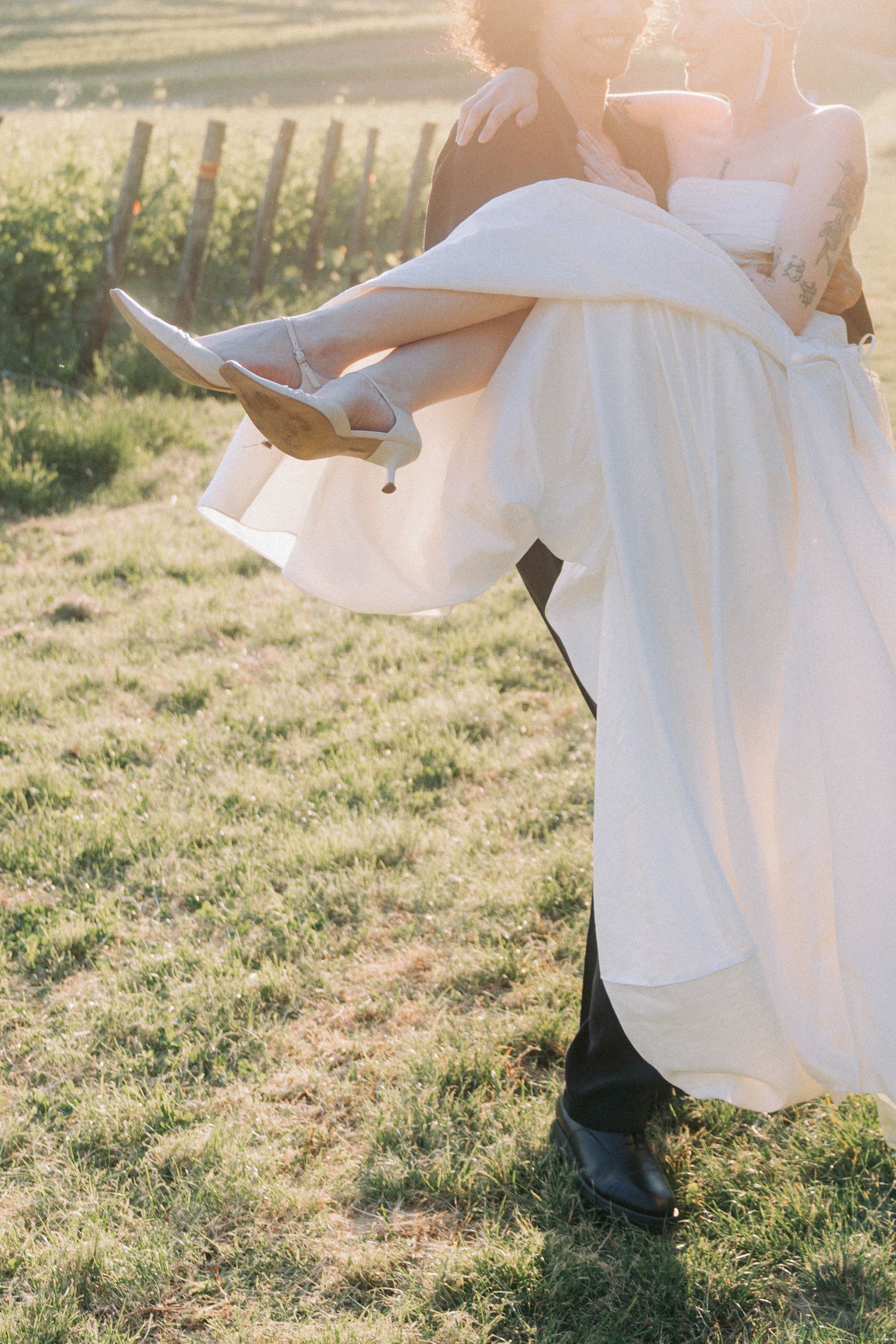Close-up view of white wedding dress and black shoes in sunlit grass during an outdoor celebration.