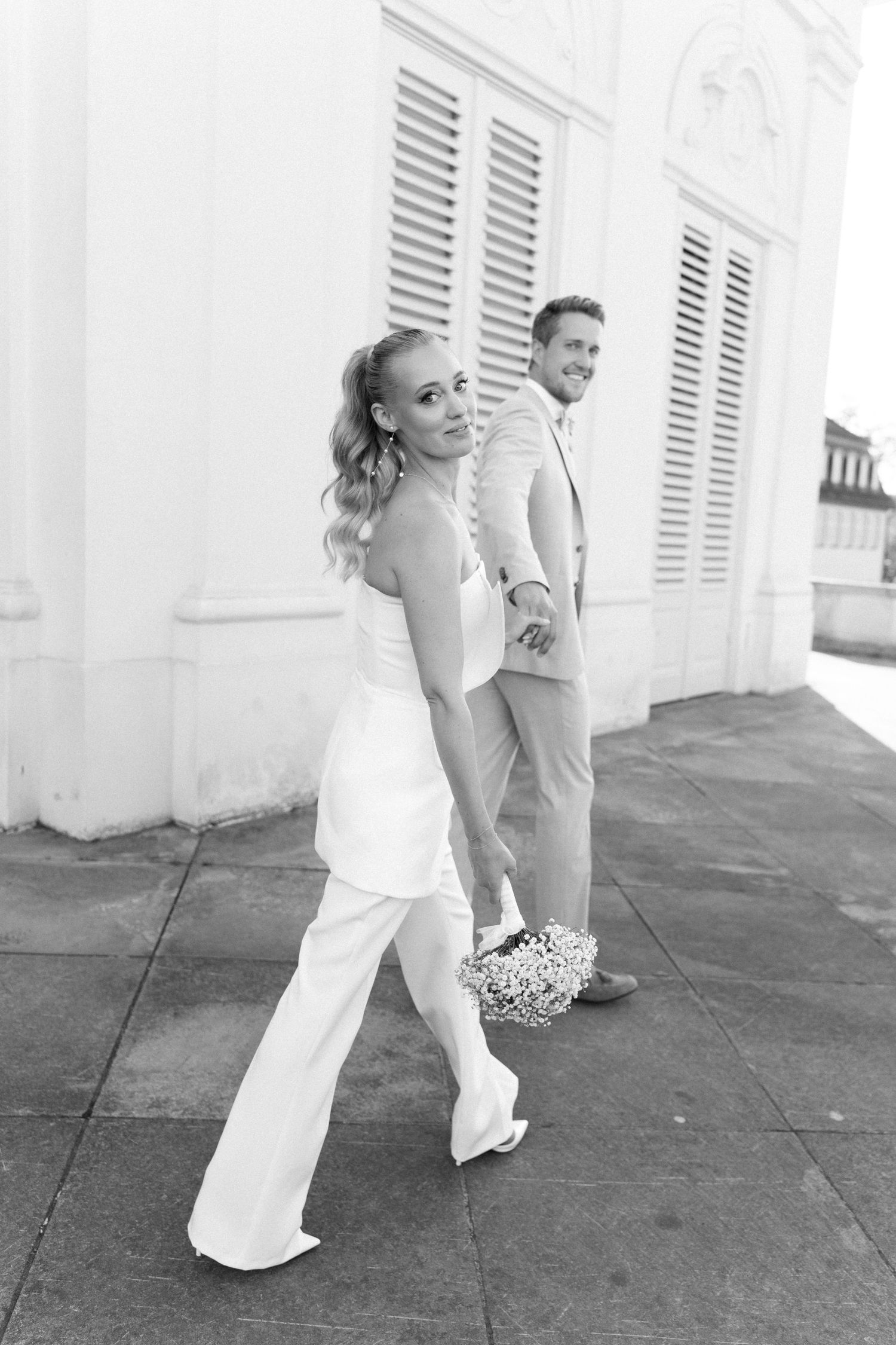 A couple in white wedding attire poses playfully outside a building with white shutters in black and white photography. Schloss Solitude