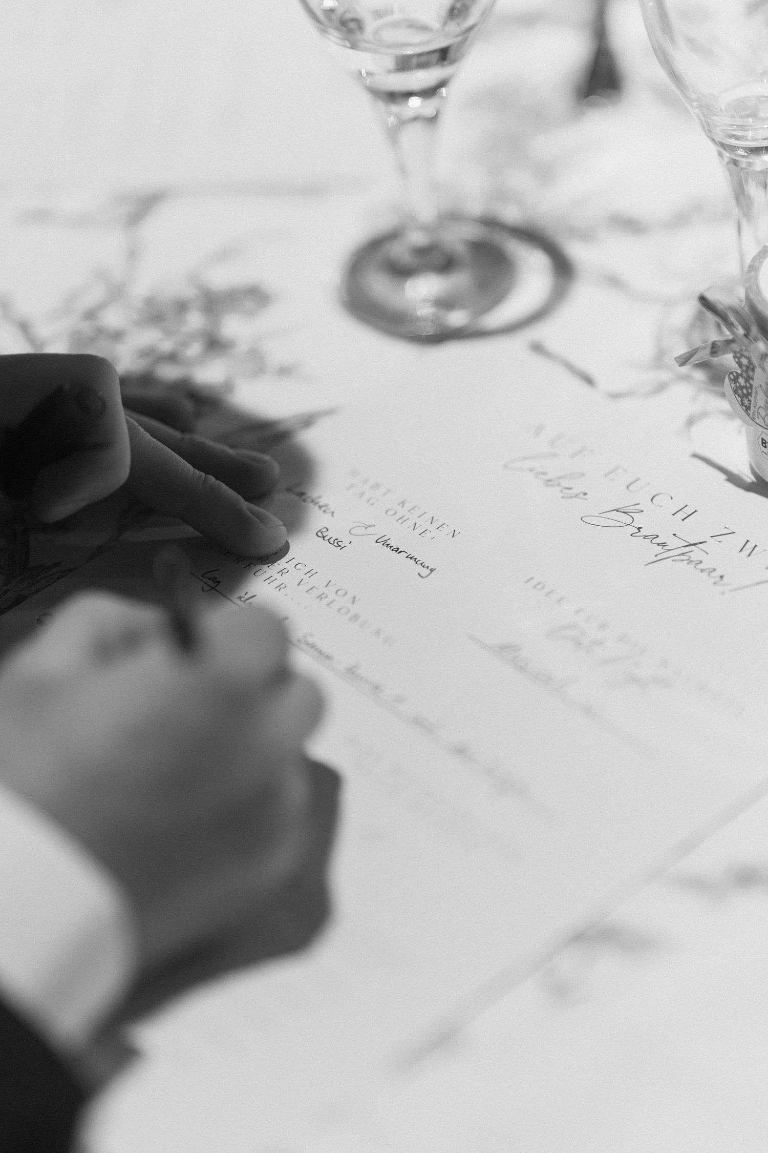 Black and white close-up of someone signing a document with wine glasses and roses in soft focus background.