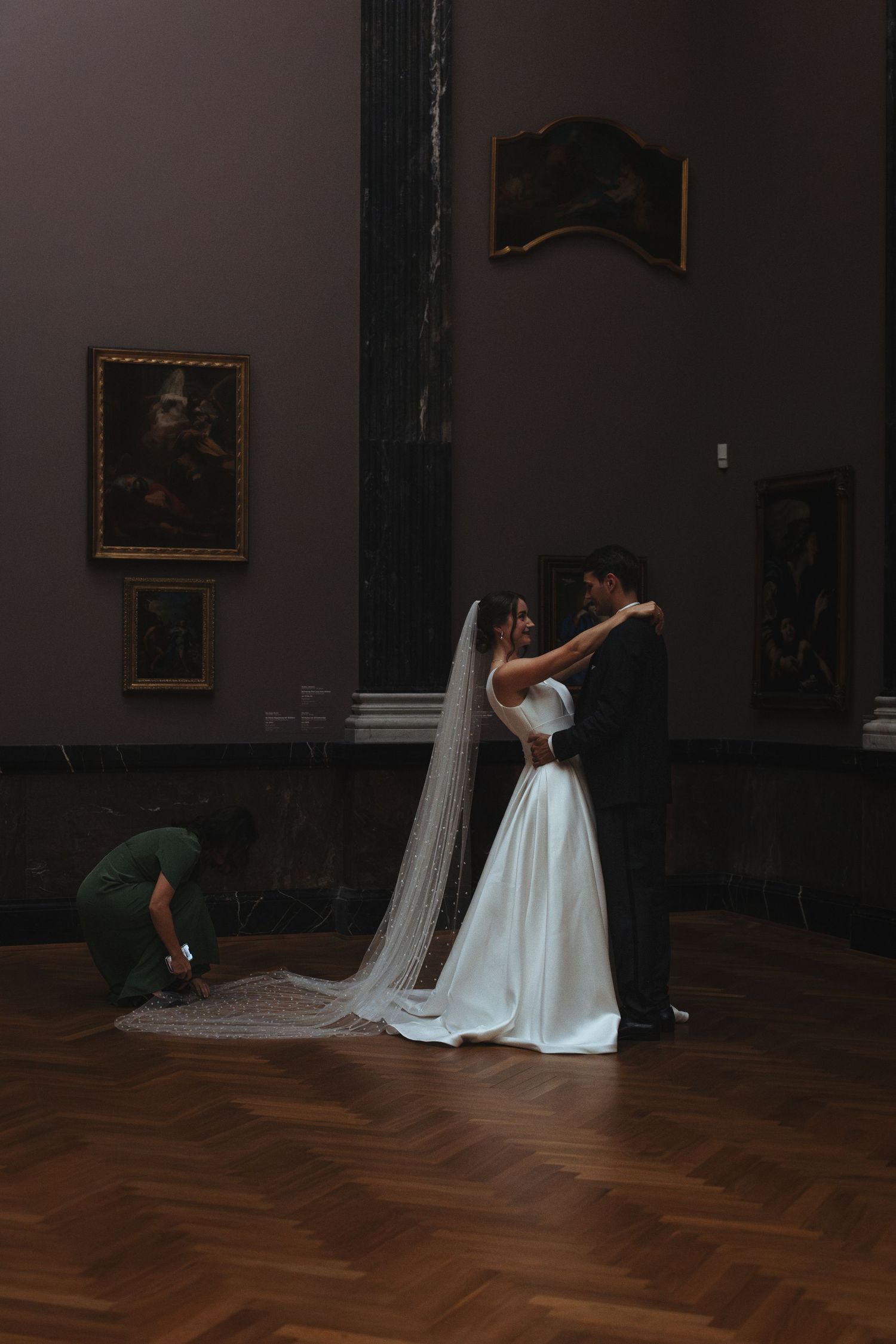A bride in a white wedding gown with a long flowing veil poses dramatically in a dark museum gallery with hardwood floors.