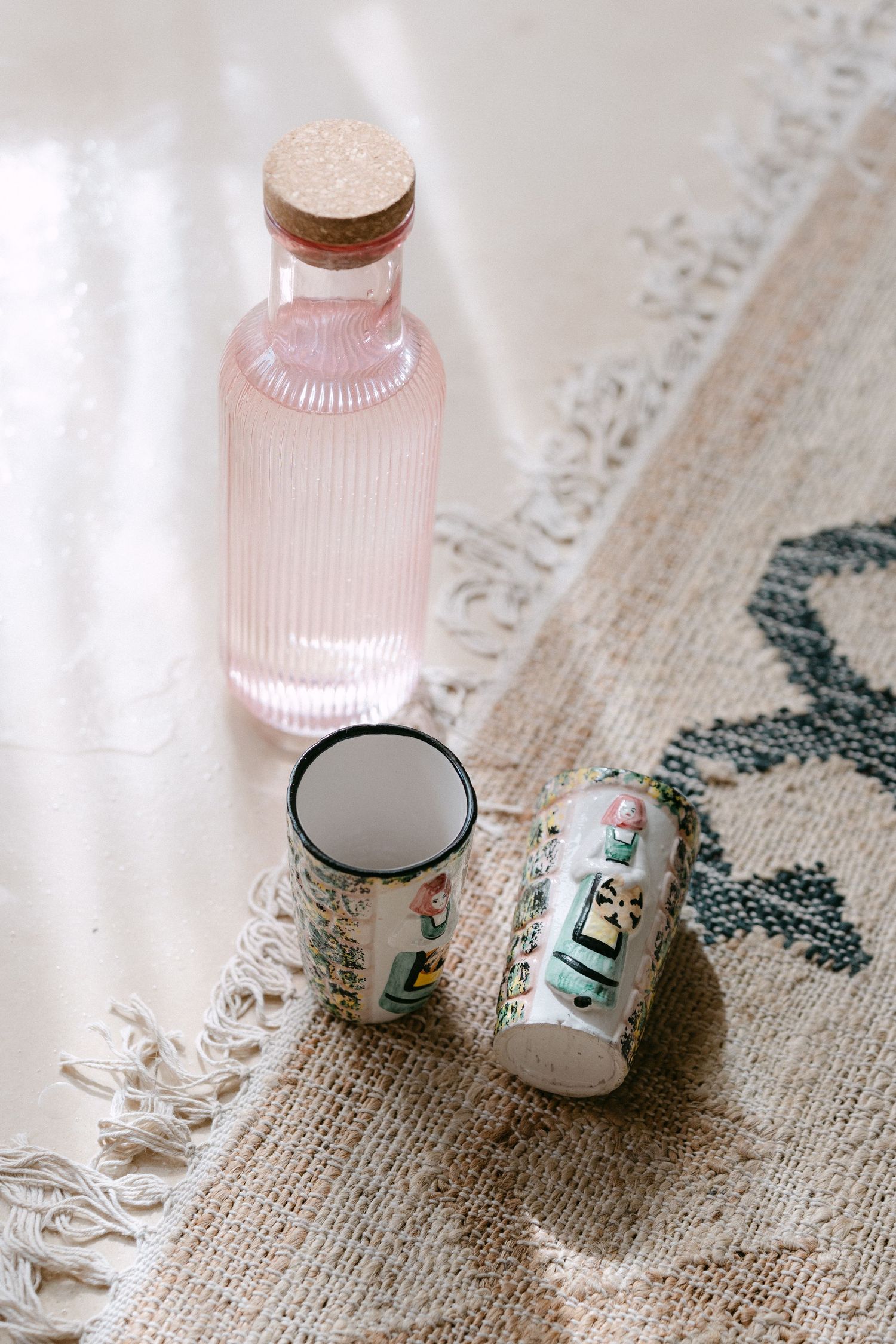 Pink glass bottle and small enamel cups arranged on a decorative patterned surface.