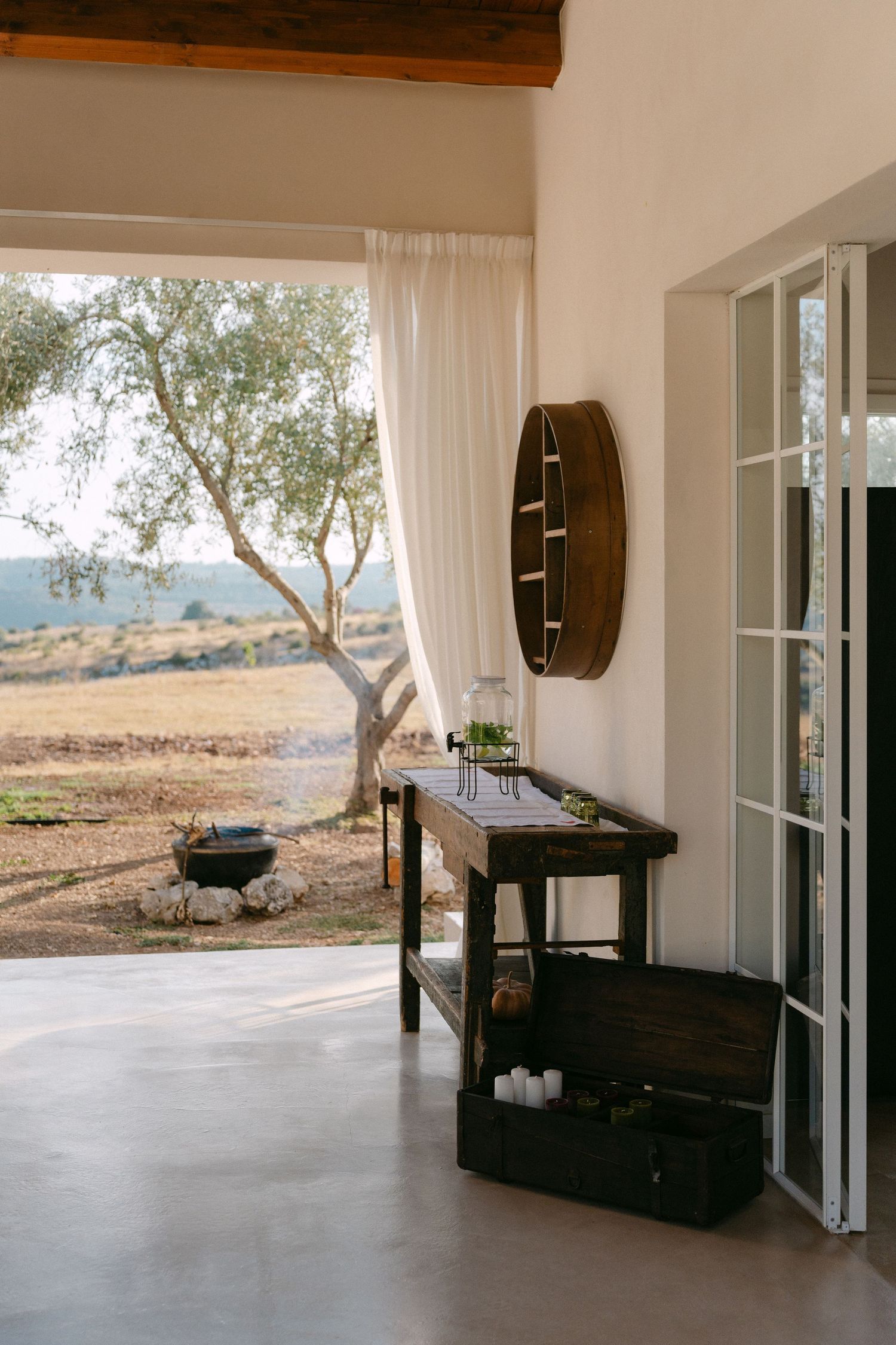 Rustic outdoor living space with wooden console table and round mirror facing olive trees and distant hills.
