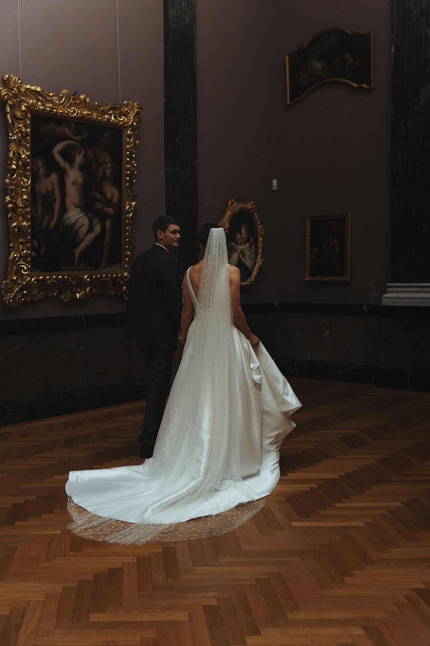 A bride in an elegant white wedding dress with a long train stands in a museum gallery with ornate gold-framed artwork.