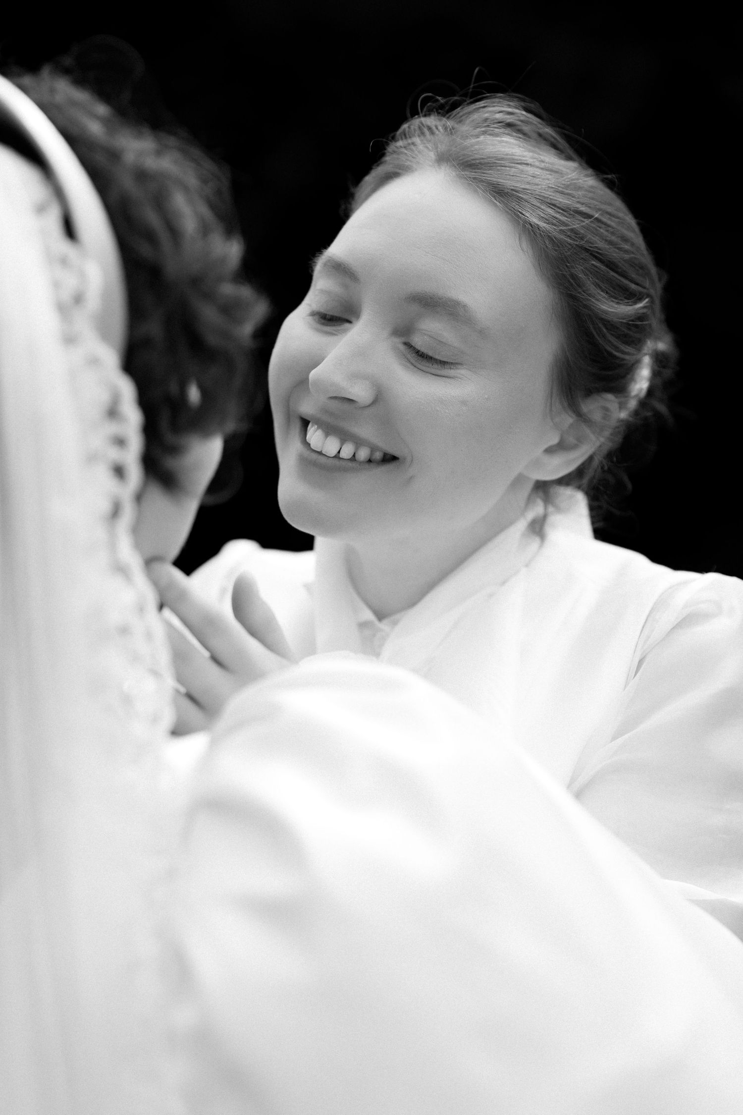 Black and white photo of a smiling bridesmaid helping a bride with her wedding dress during bridal preparations.