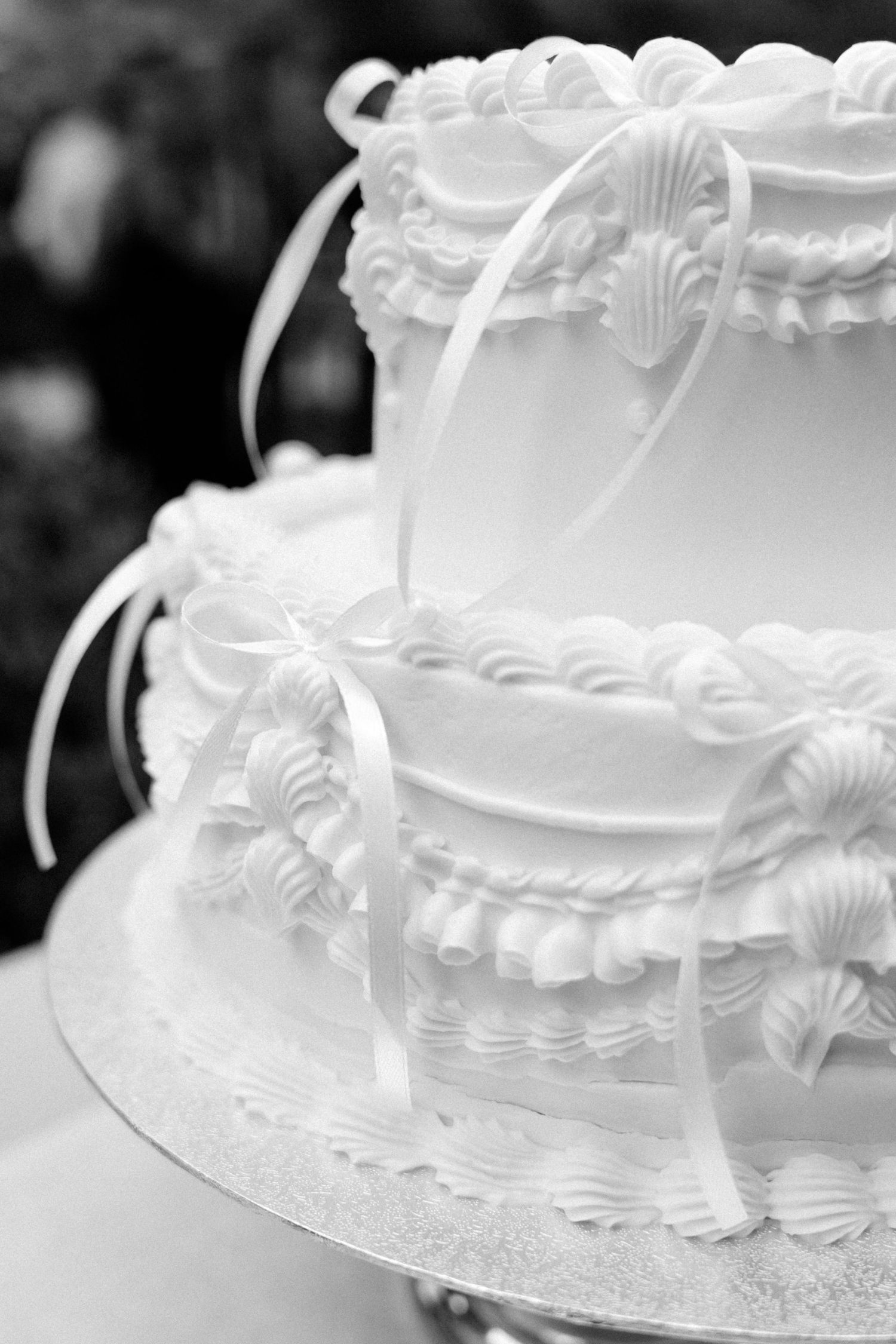 Black and white photo of a two-tier wedding cake with piped shell borders, ruffled icing details, and satin ribbons on a silver cake board.