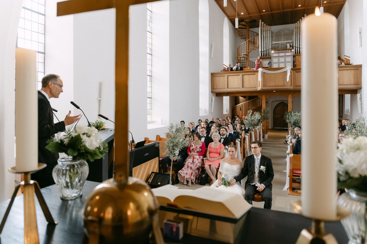 Officiant speaks at a podium during a church wedding ceremony as the bride and groom stand before seated guests, with candles and flowers in