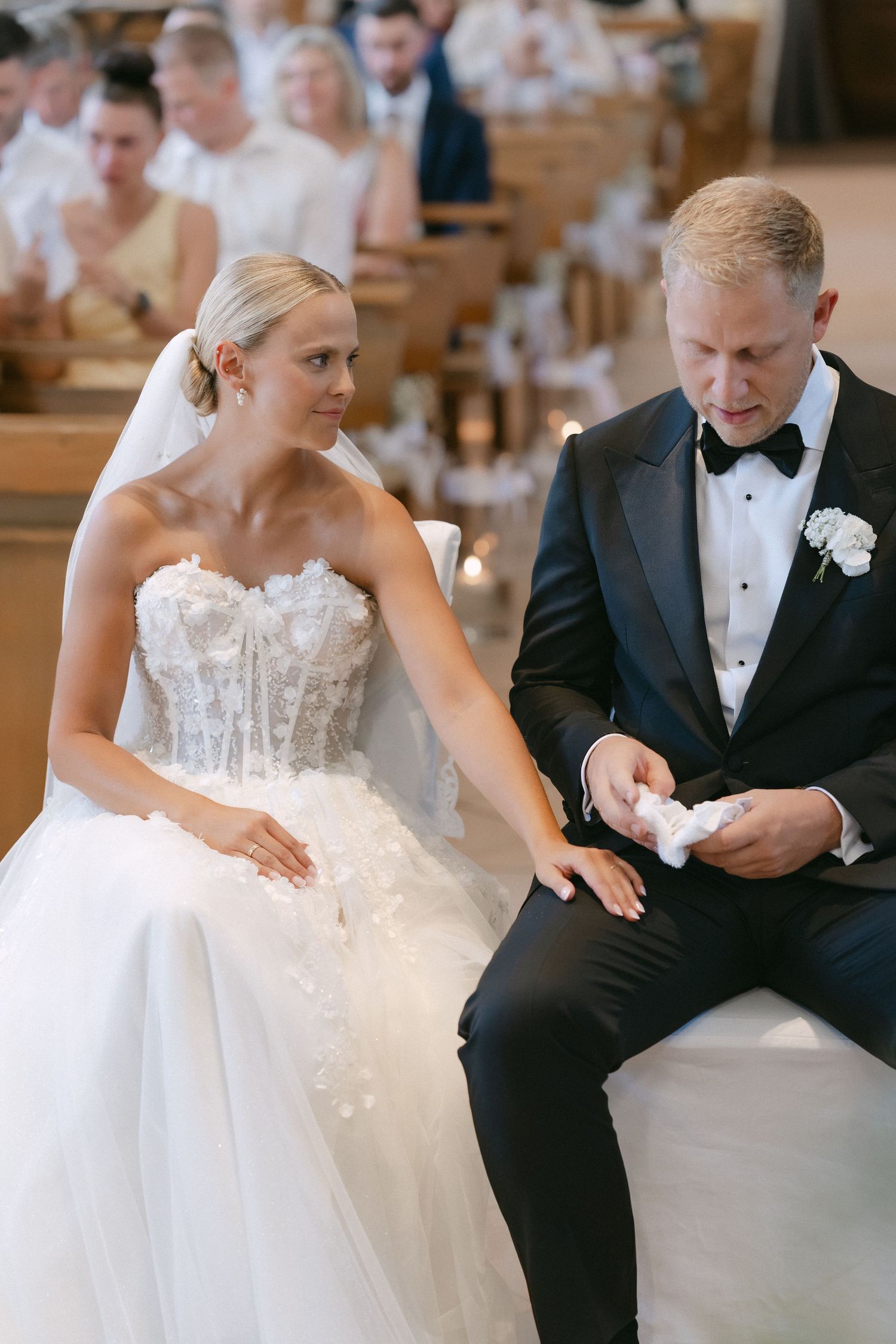 Bride in a strapless lace wedding gown sits beside a groom in a black tuxedo holding a handkerchief during a church ceremony.