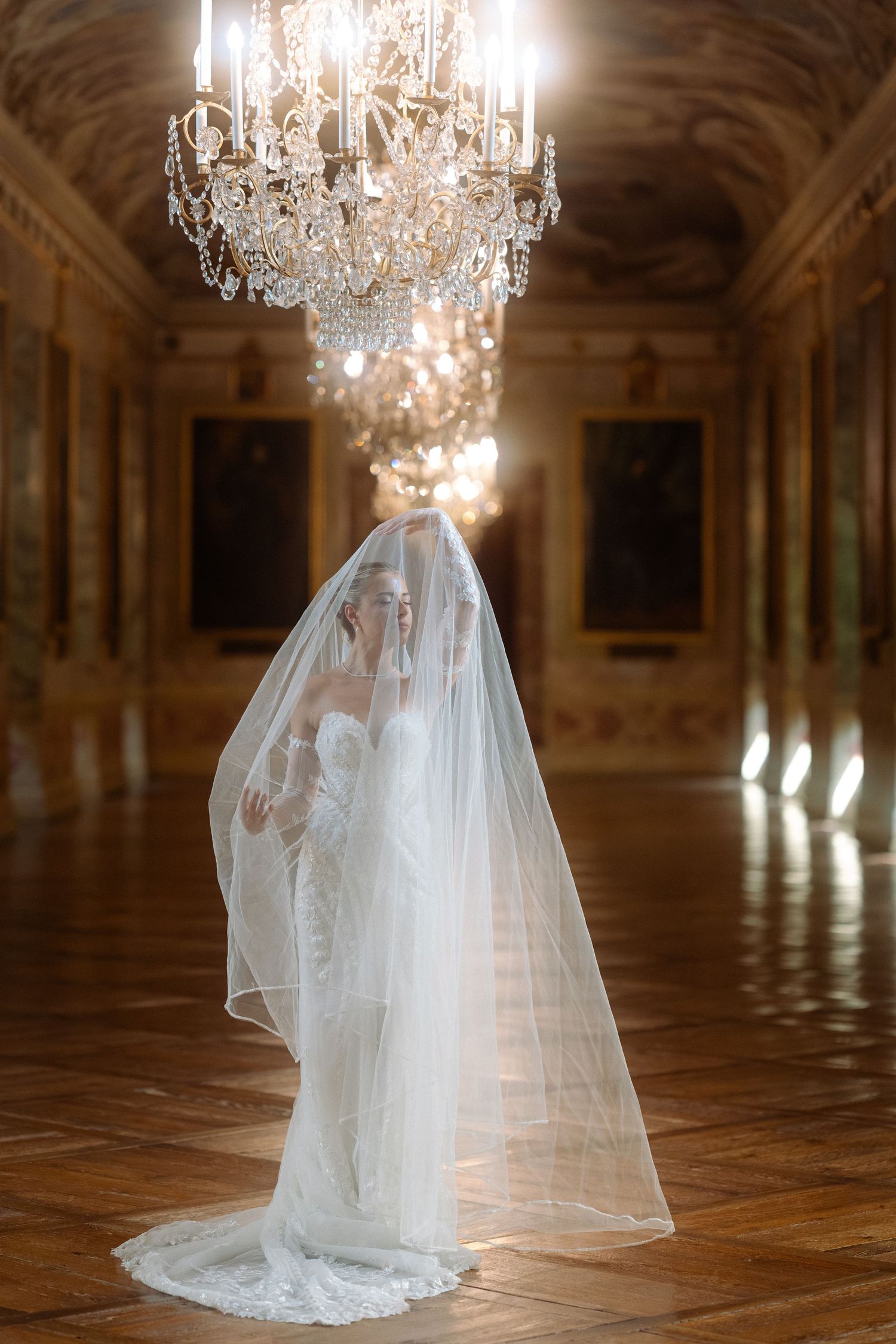 Elegant bride in flowing white gown and veil stands beneath ornate crystal chandelier in grand palace ballroom. Schloss Ludwigsburg