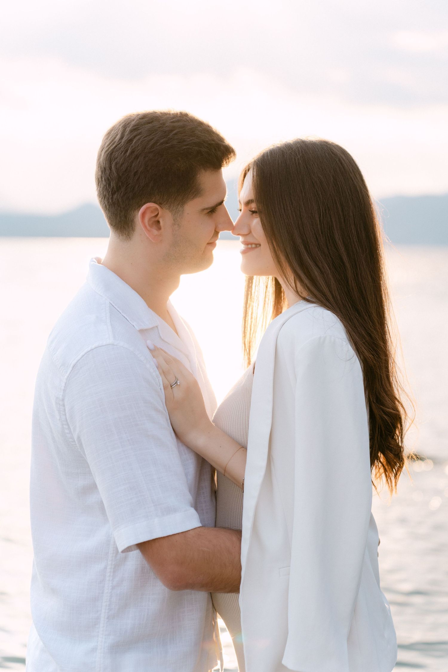 Romantic couple in white clothing embraces tenderly by the ocean at sunset.