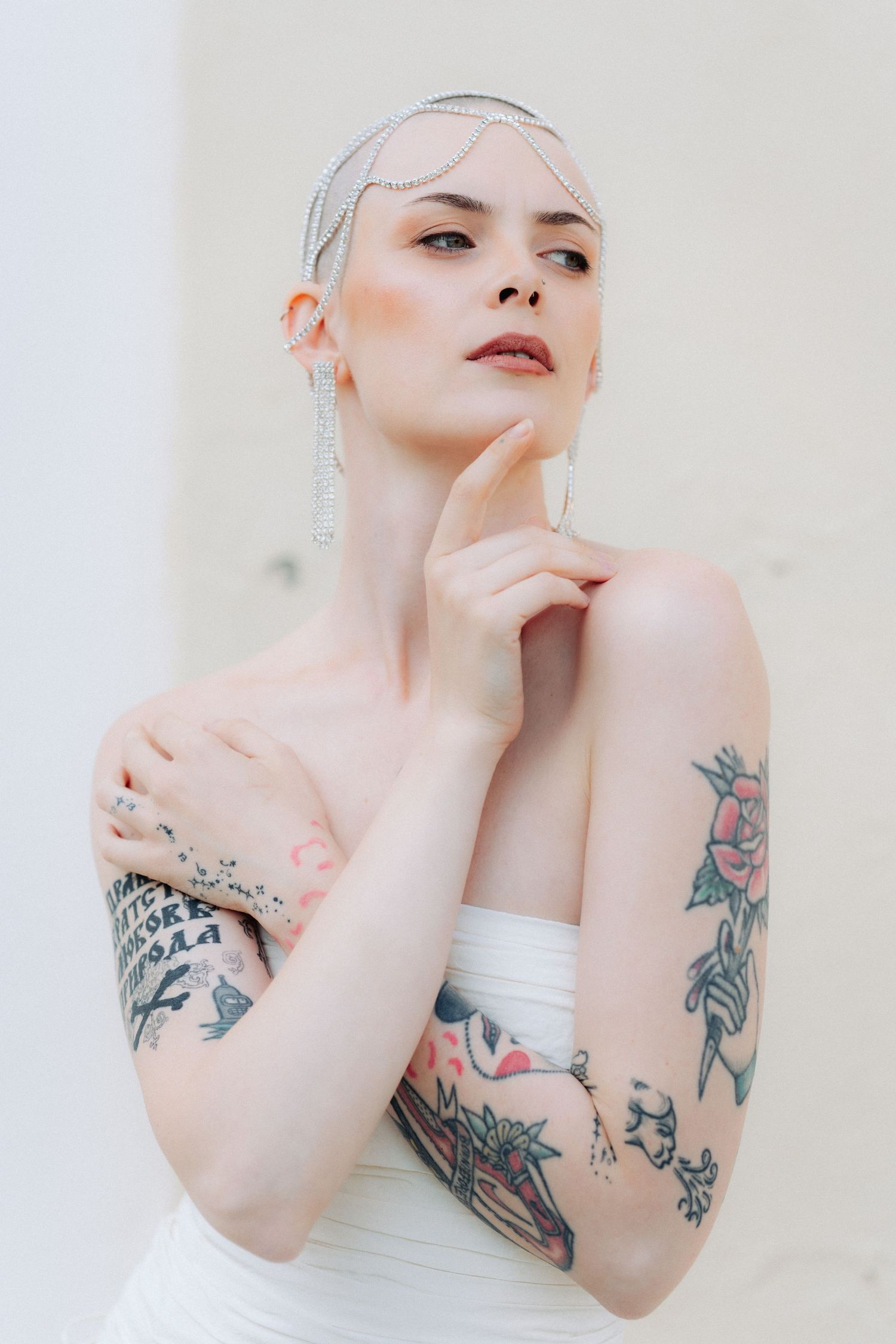 A model in white poses artistically against a light background, showing off decorative arm tattoos.
