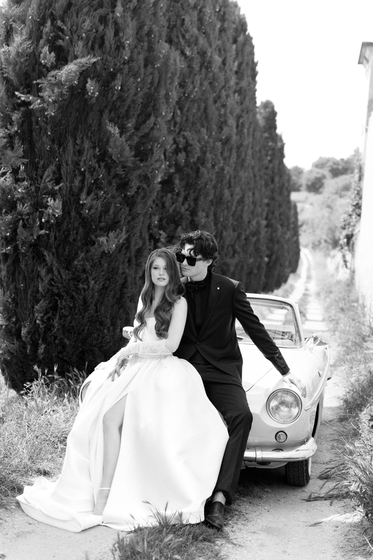 A couple in wedding attire poses next to a vintage Volkswagen Beetle on a cypress tree-lined path in black and white.