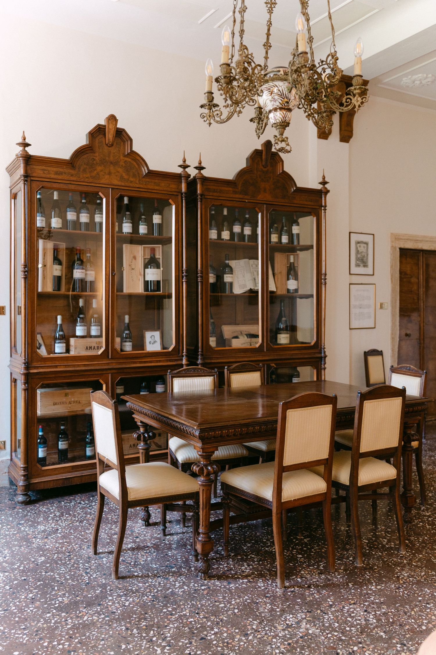 Elegant vintage dining room with ornate wooden display cabinets, upholstered chairs, and brass chandelier on terrazzo floor. Villa Mosconi