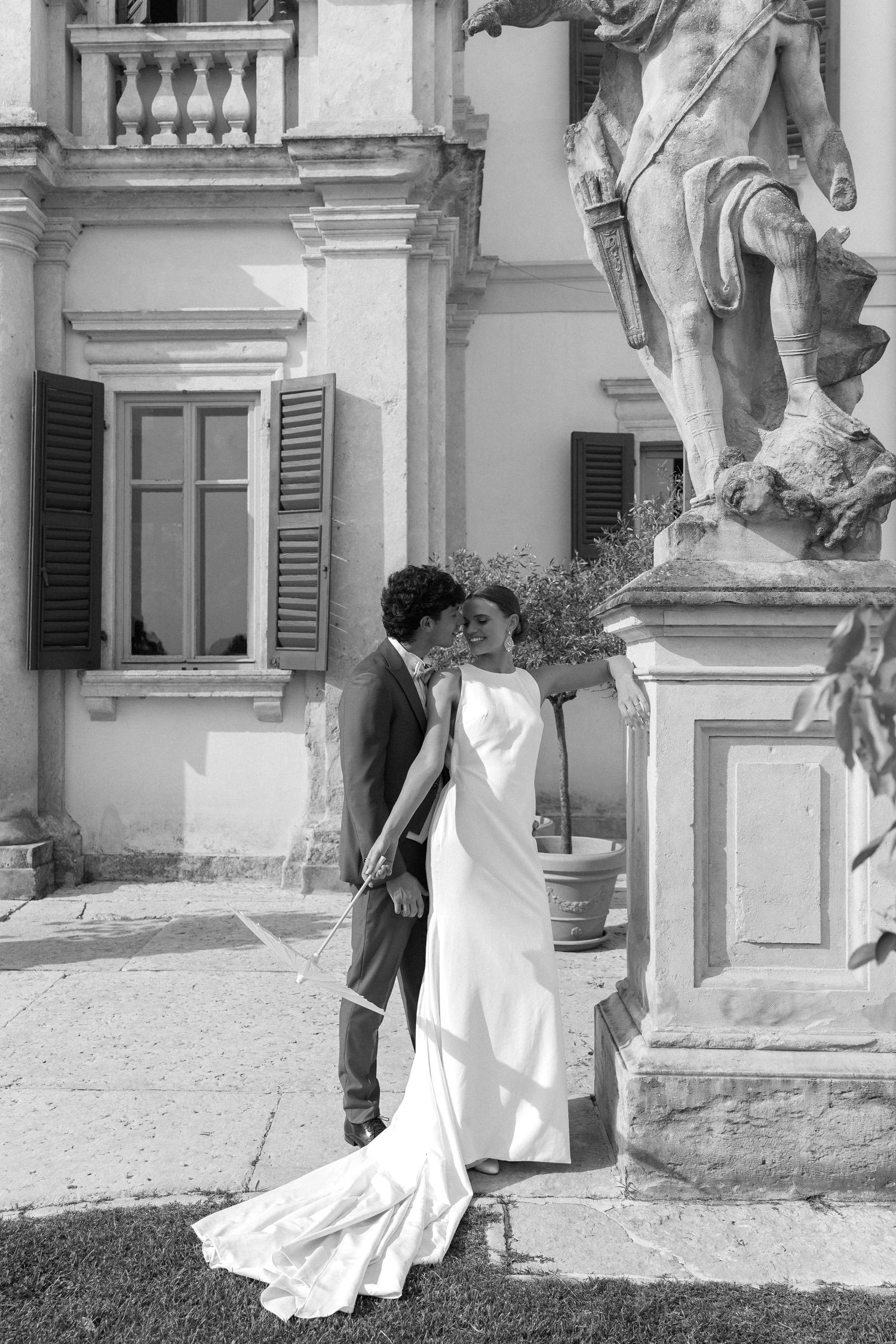 A romantic black and white wedding photo of a couple embracing by an elegant Italian villa with classic architecture. Villa Mosconi Bertani