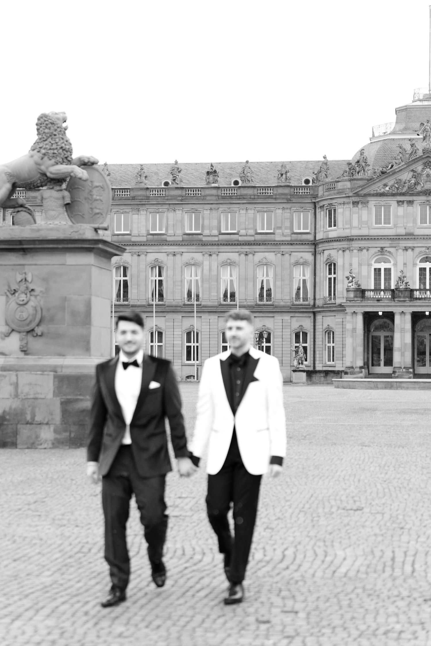 A black and white photo of two people in tuxedos walking together outside an elegant European palace building.