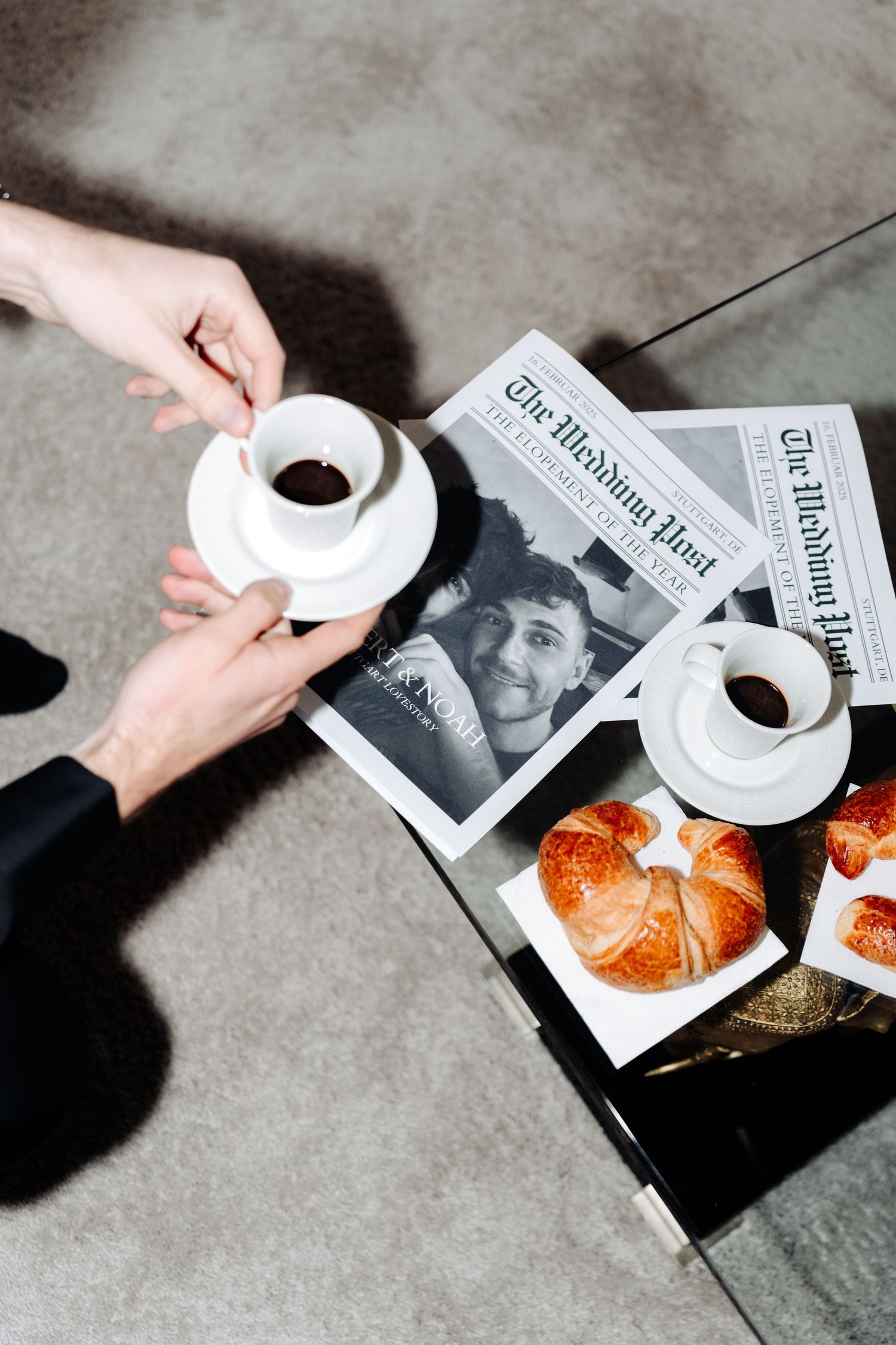 Overhead view of hands passing coffee cups with newspaper and croissants on concrete surface.