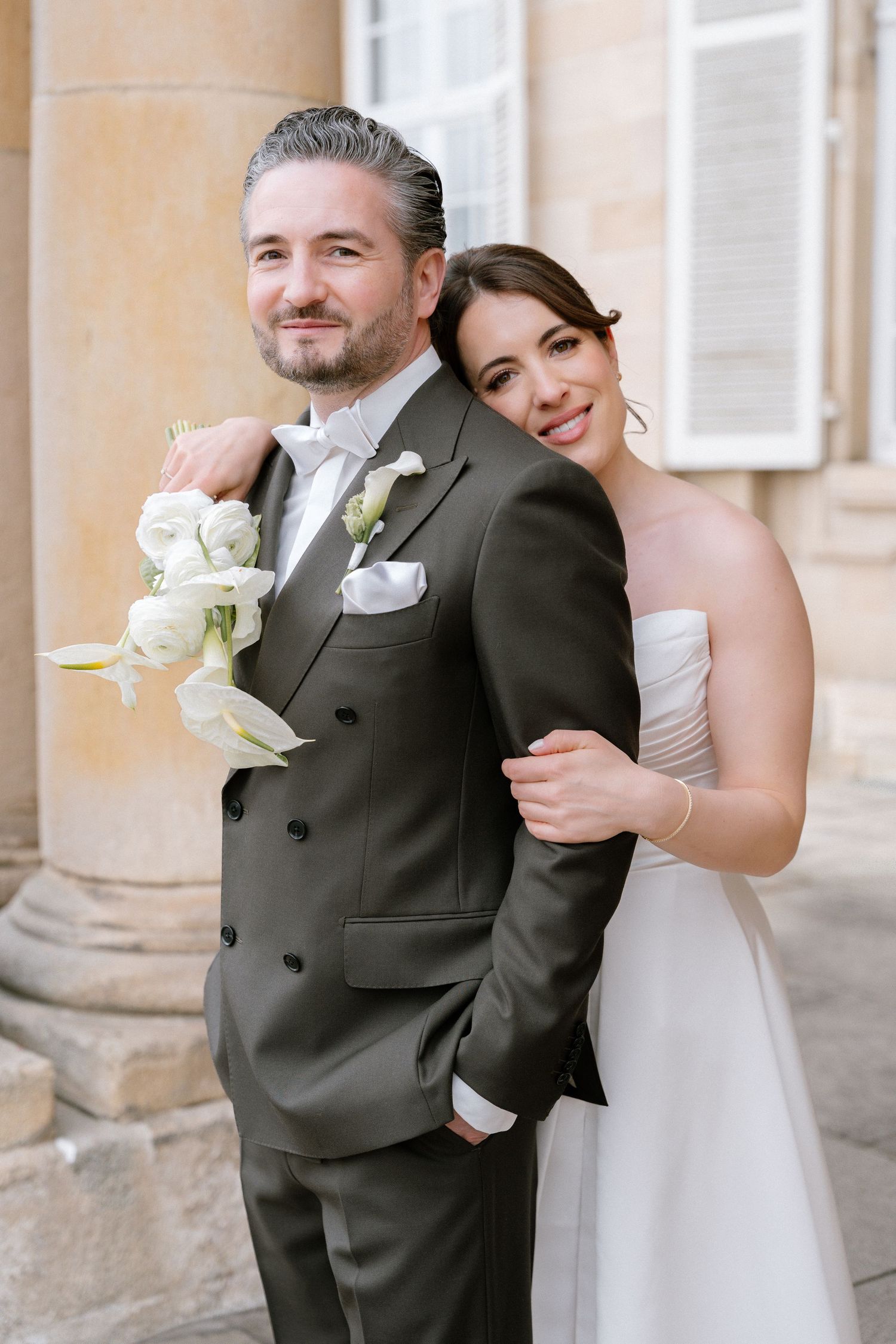 A smiling bride in a white strapless gown embraces a groom in a dark suit holding white flowers outdoors.