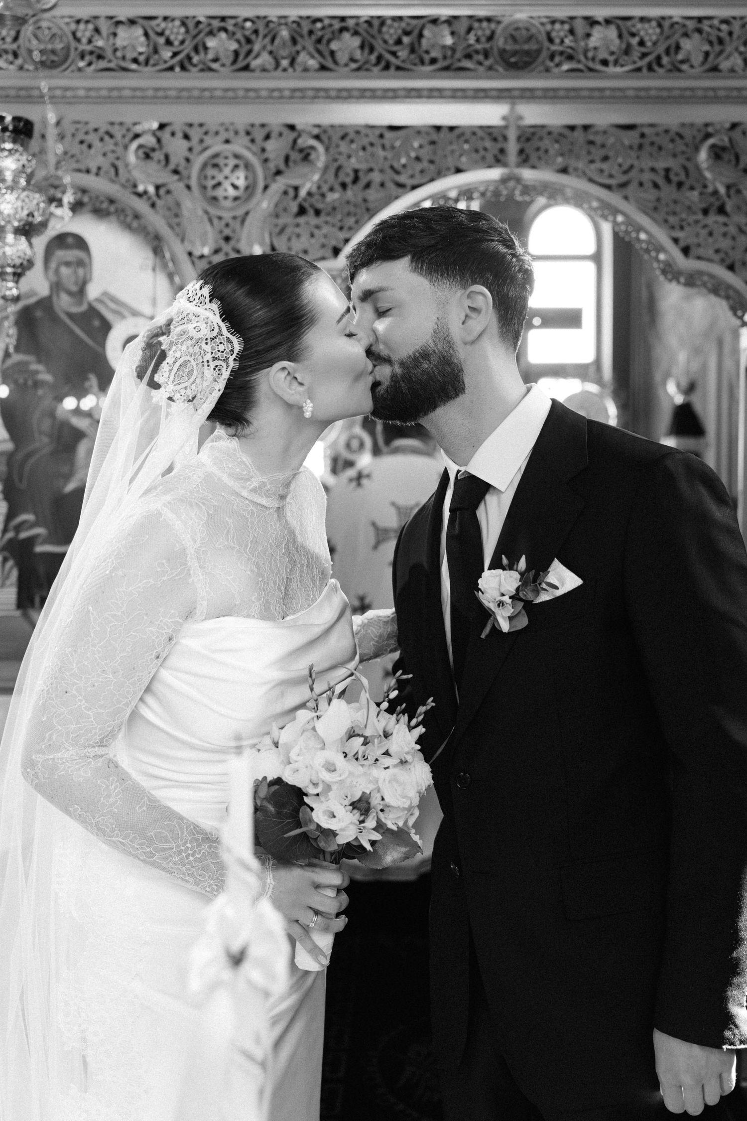 Black and white photo of a bride and groom sharing a kiss during their wedding ceremony inside an ornate church.