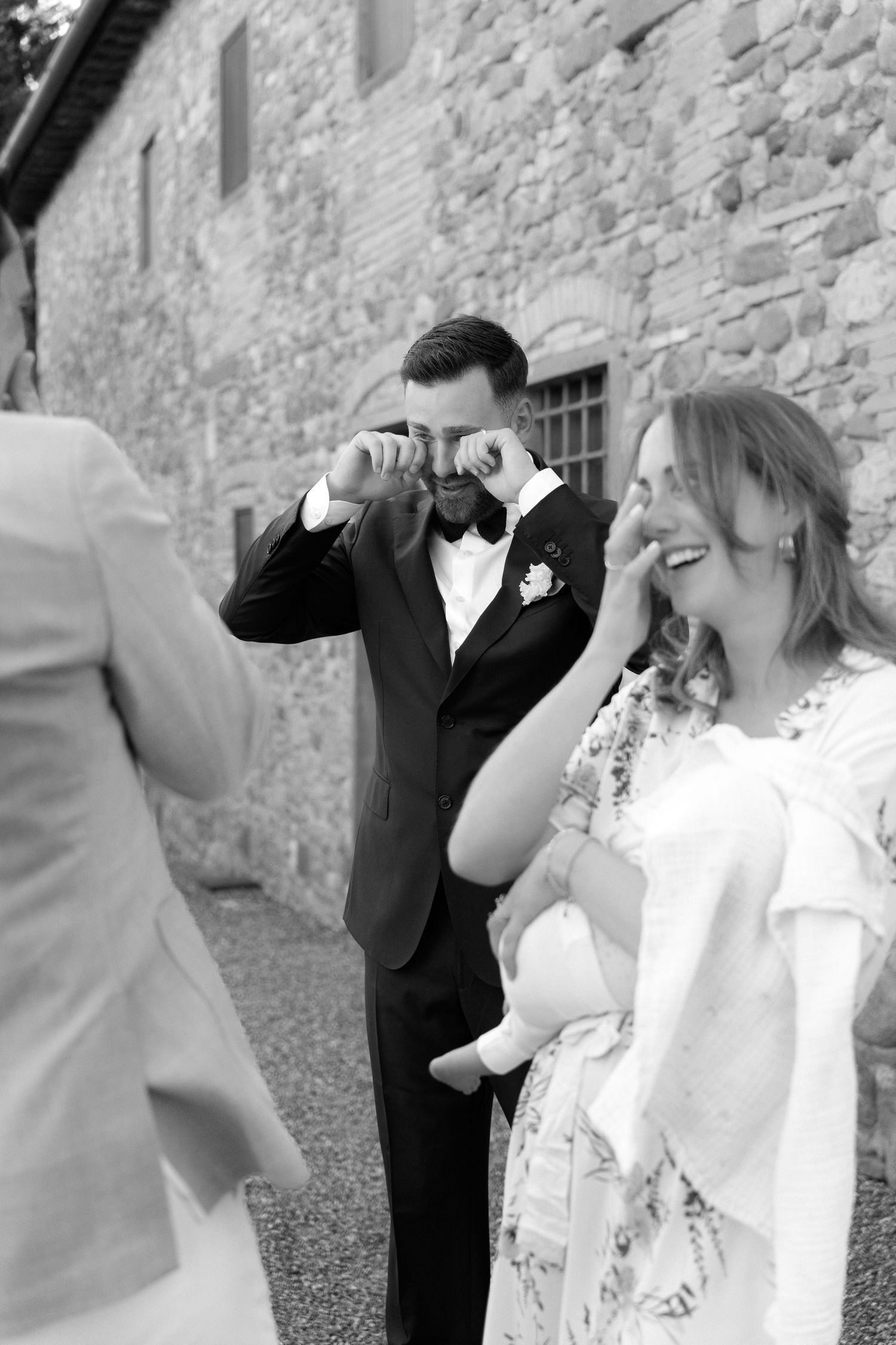 Groom wiping tears of joy at wedding while guest laughs beside him, black and white photo at rustic stone venue.