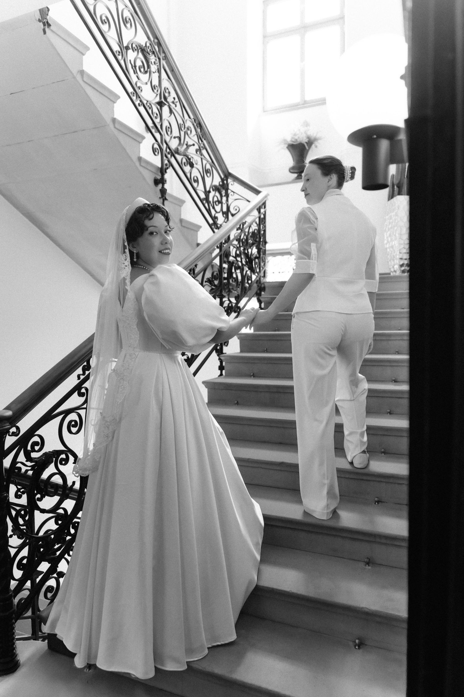 Black and white photo of a bride in a white gown smiling on elegant stairs with a partner ascending ahead.