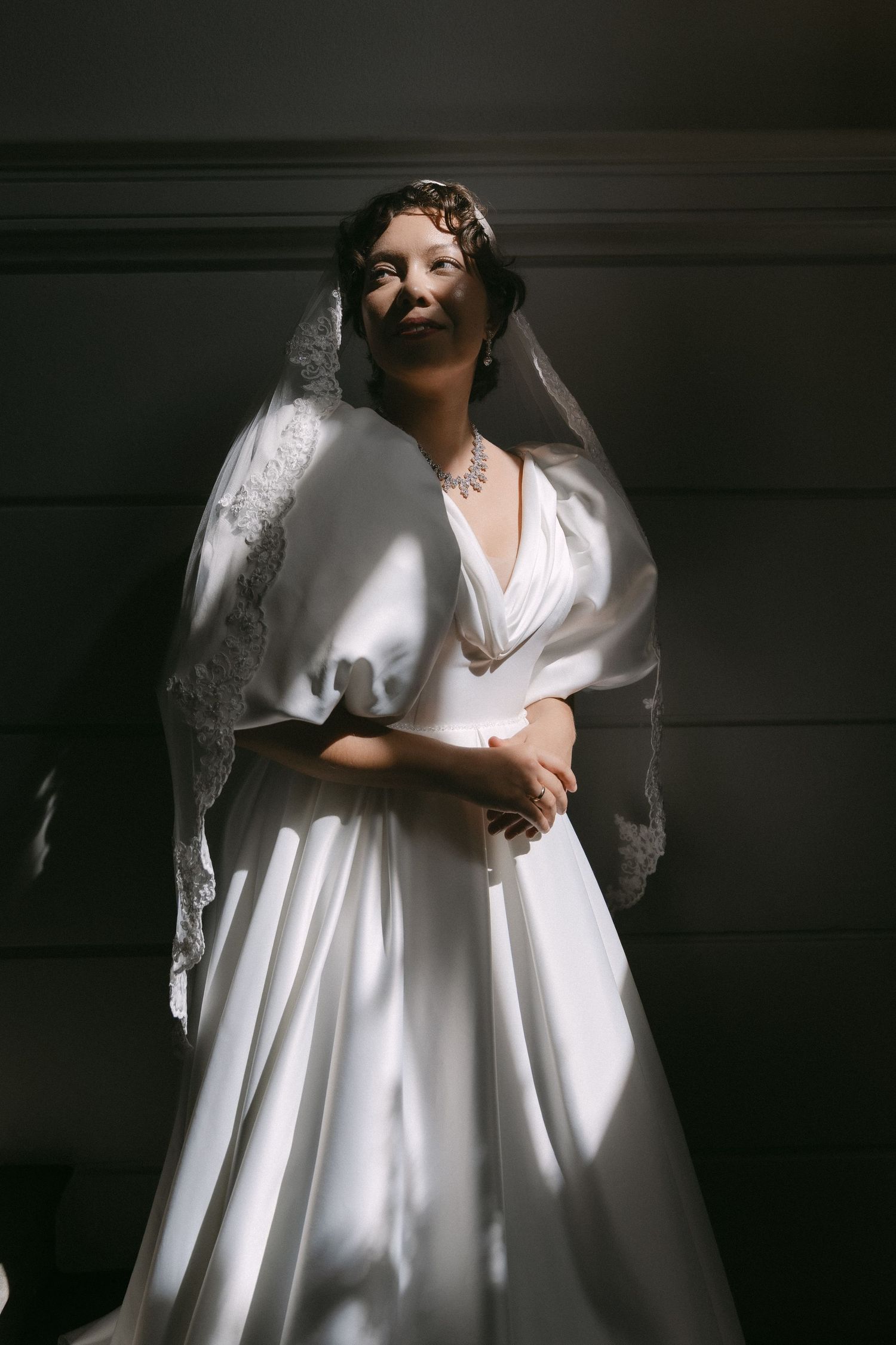 Bride in white satin gown with lace veil and diamond necklace poses in dramatic natural light against dark wall.